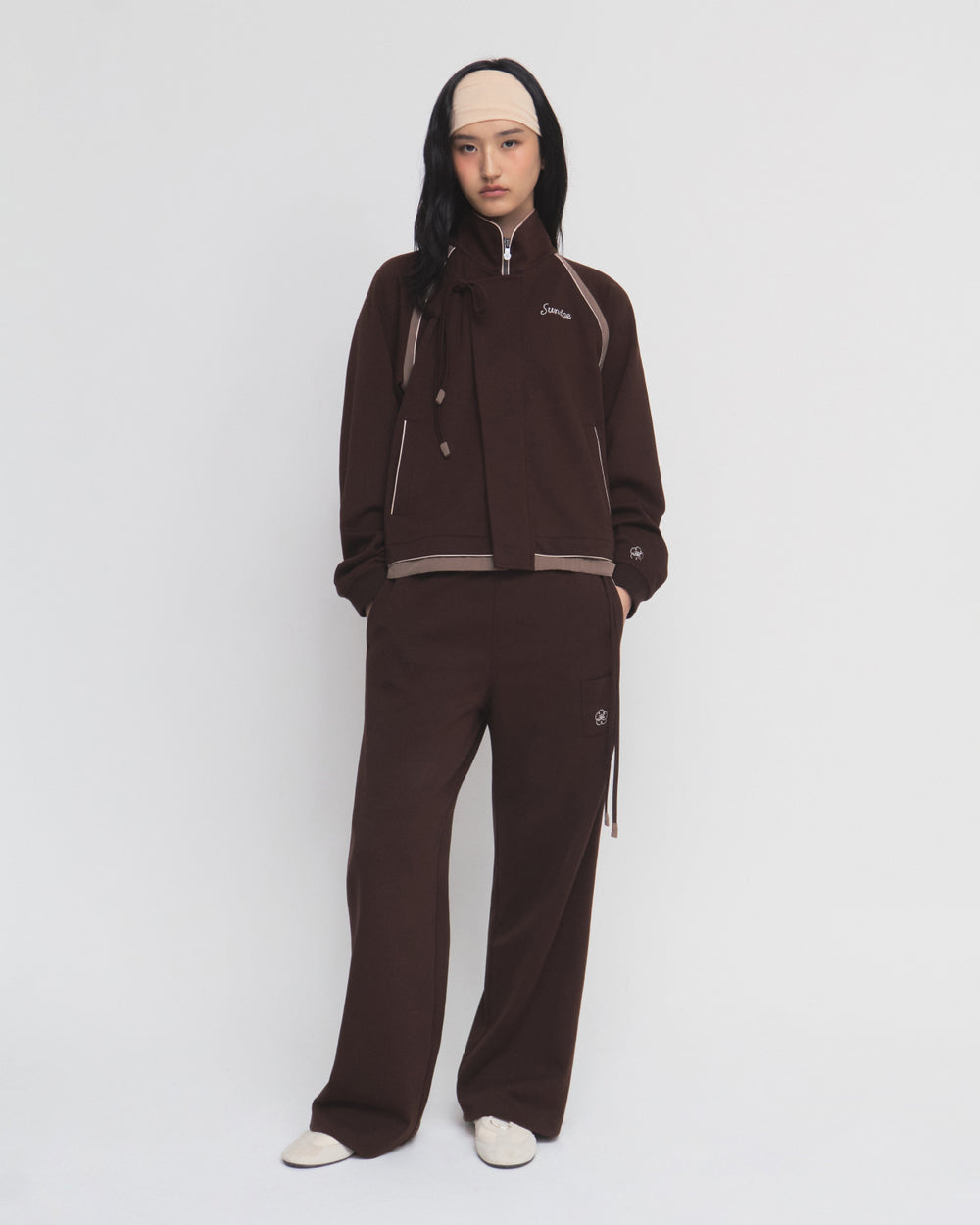 A woman stands against a plain white background wearing the Sundae School BROWN PALISADE PARK TRACK JACKET with contrast fabric details, matching pants, white shoes, and a light beige headband.