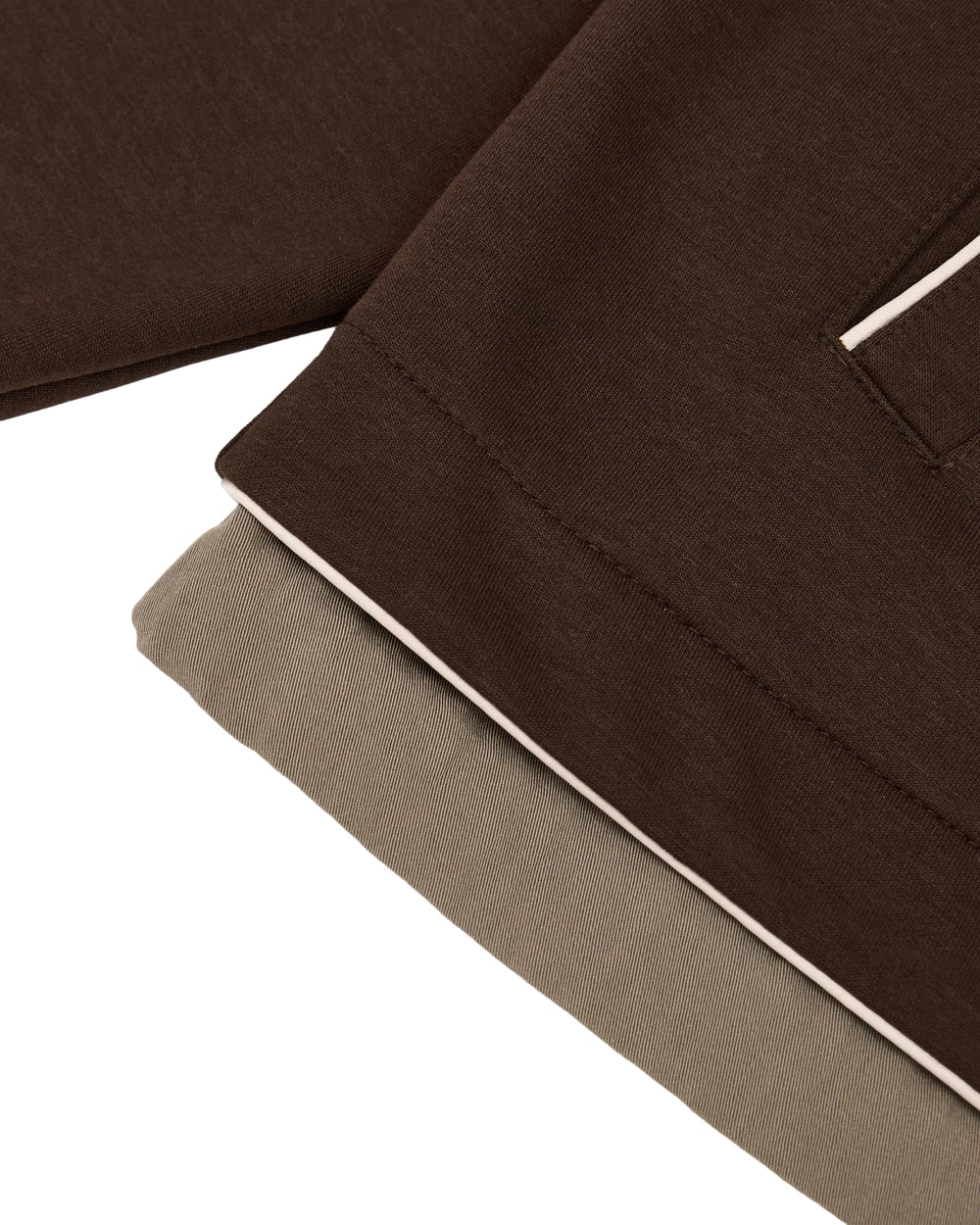 Close-up of overlapping brown and beige fabric from the Sundae School BROWN PALISADE PARK TRACK JACKET, showcasing visible stitching and a pocket edge finished with a thin, light-colored contrast trim—details evocative of classic athletic jackets.