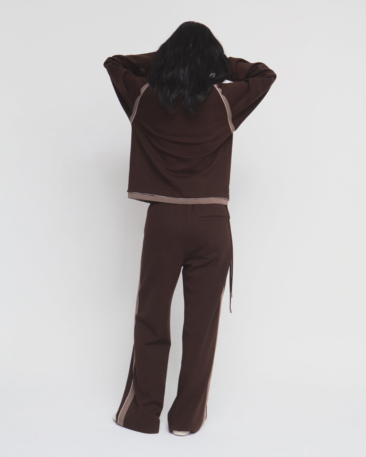 A person with long dark hair, seen from behind, is wearing the Sundae School BROWN PALISADE PARK TRACK JACKET, featuring light beige side stripes and contrast fabric details, while standing against a plain white background.