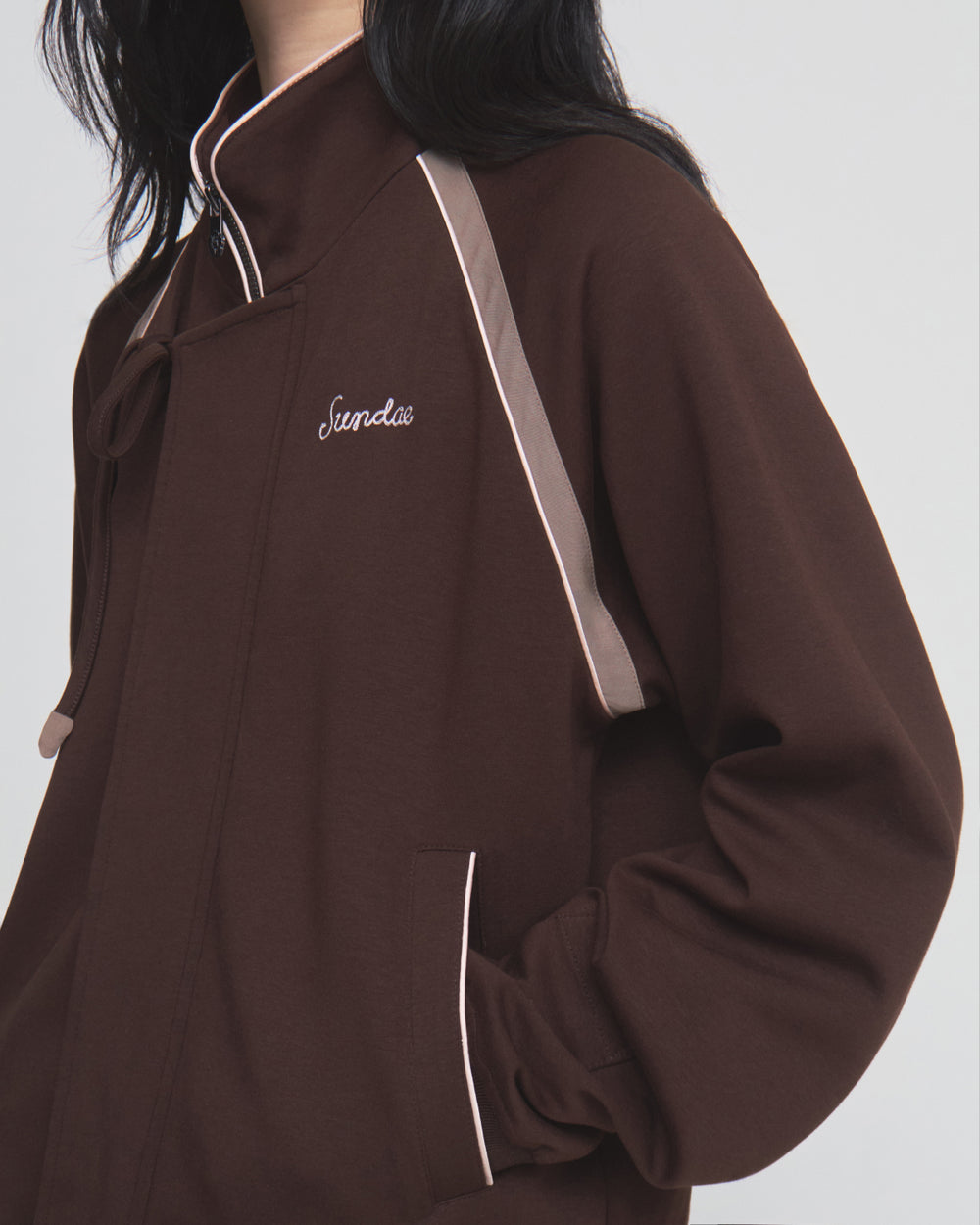 A person wears the Sundae School BROWN PALISADE PARK TRACK JACKET, featuring light pink trim and “Sunday” chain-stitch embroidery on the chest. The person’s face is not visible, and their hand is in the pocket.