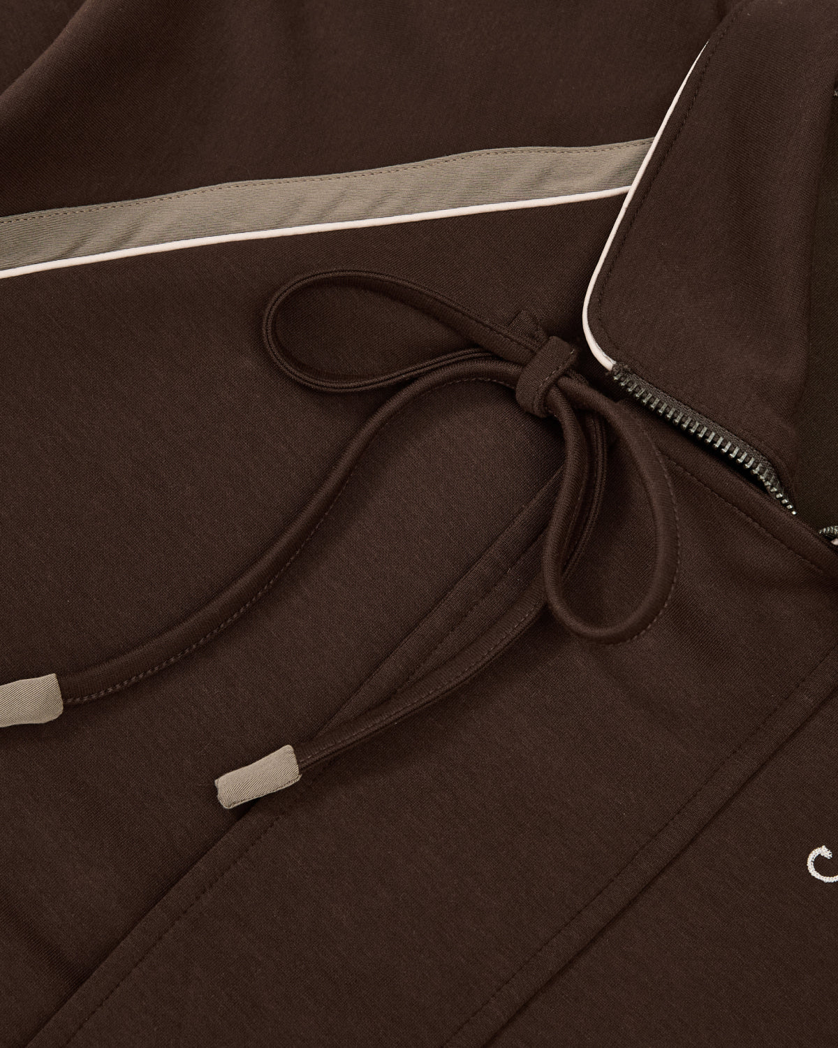 Close-up of the Sundae School BROWN PALISADE PARK TRACK JACKET, featuring a brown athletic design with a slim white stripe and taupe accent. The visible drawstrings are tied in a bow, while the partially open zipper reveals a glimpse of the inner collar. Subtle contrast fabric details elevate its versatile style.