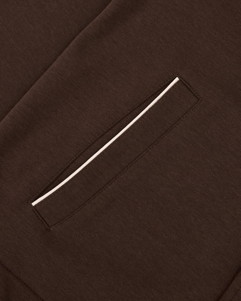 Close-up of the BROWN PALISADE PARK TRACK JACKET by Sundae School showcases a brown fabric surface featuring a welt pocket with a thin white trim along the edge. Visible stitching highlights the garment’s craftsmanship, echoing the precision typical of an athletic track jacket, while subtle contrast fabric details enhance its contemporary style.