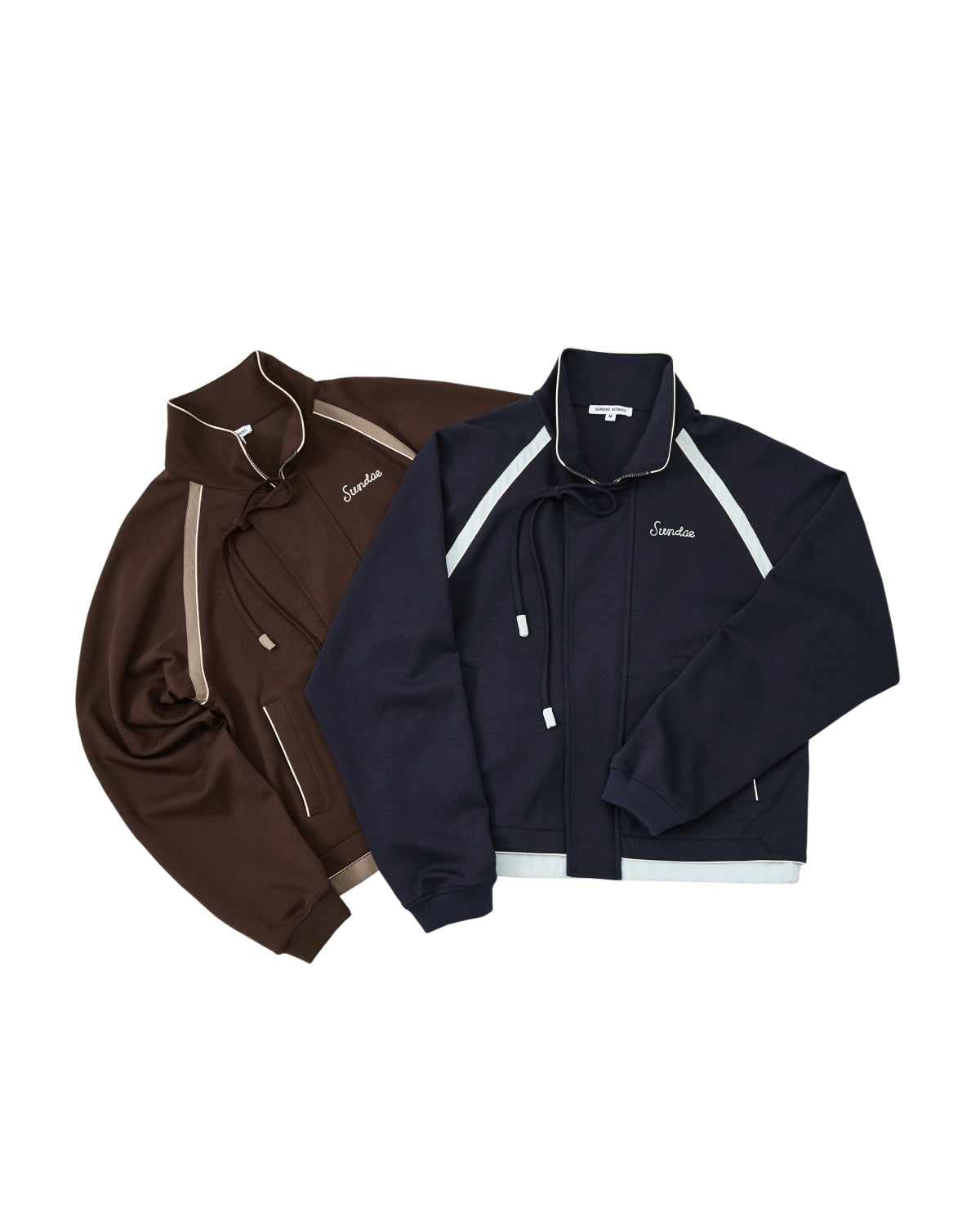 Two zip-up Sundae School BROWN PALISADE PARK TRACK JACKETS are laid flat—one in brown with beige contrast fabric accents, and the other in navy blue with white accents—both showcasing white Emelen chain-stitch embroidery on the chest for a classic athletic track jacket look.