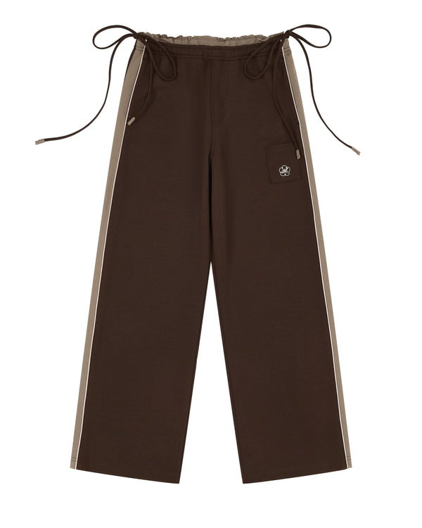 The Sundae School BROWN PALISADE PARK TRACK PANTS are wide-leg boot cut track pants in brown, designed with beige contrast piping along the sides. They feature an elastic waistband with long drawstrings and a front patch pocket embellished with a small embroidered skull, shown laid flat on a white background.