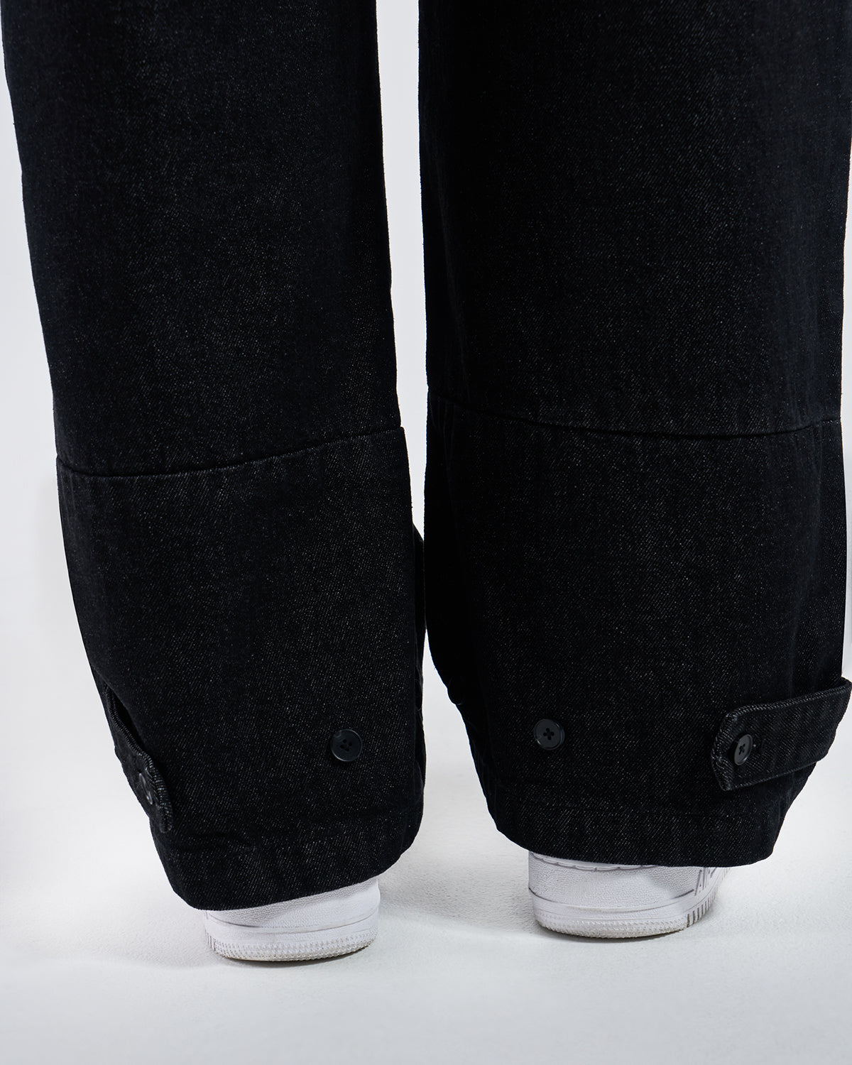 Close-up detail of black hanbok denim hem showing gathered elastic ankle cuffs with black snap button closures, modeled with white sneakers, demonstrating adjustable hem construction