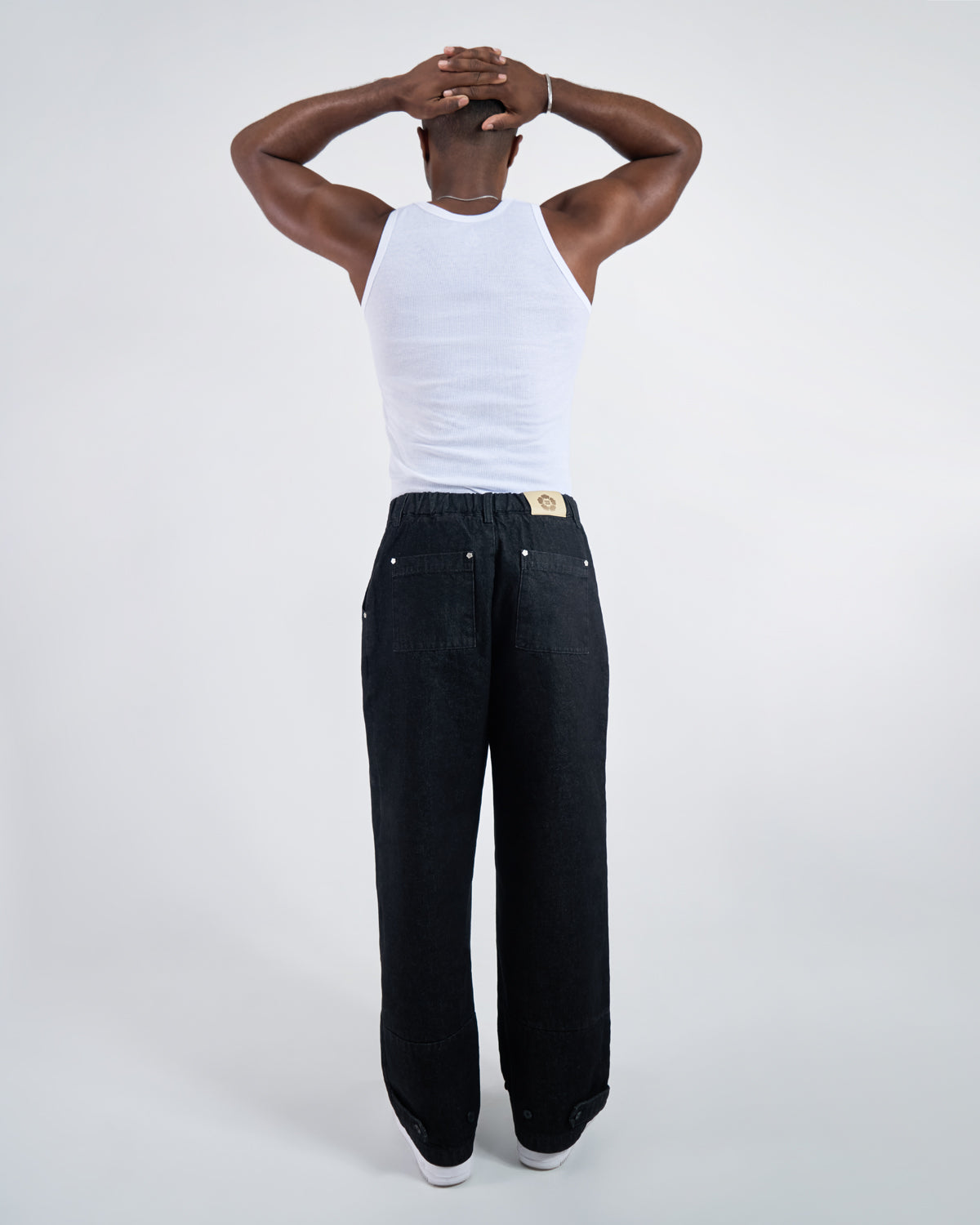 Back view of black wide-leg hanbok denim pants modeled with white ribbed tank top, hands clasped behind head pose, showing Sundae School leather patch and back pocket details against white studio background