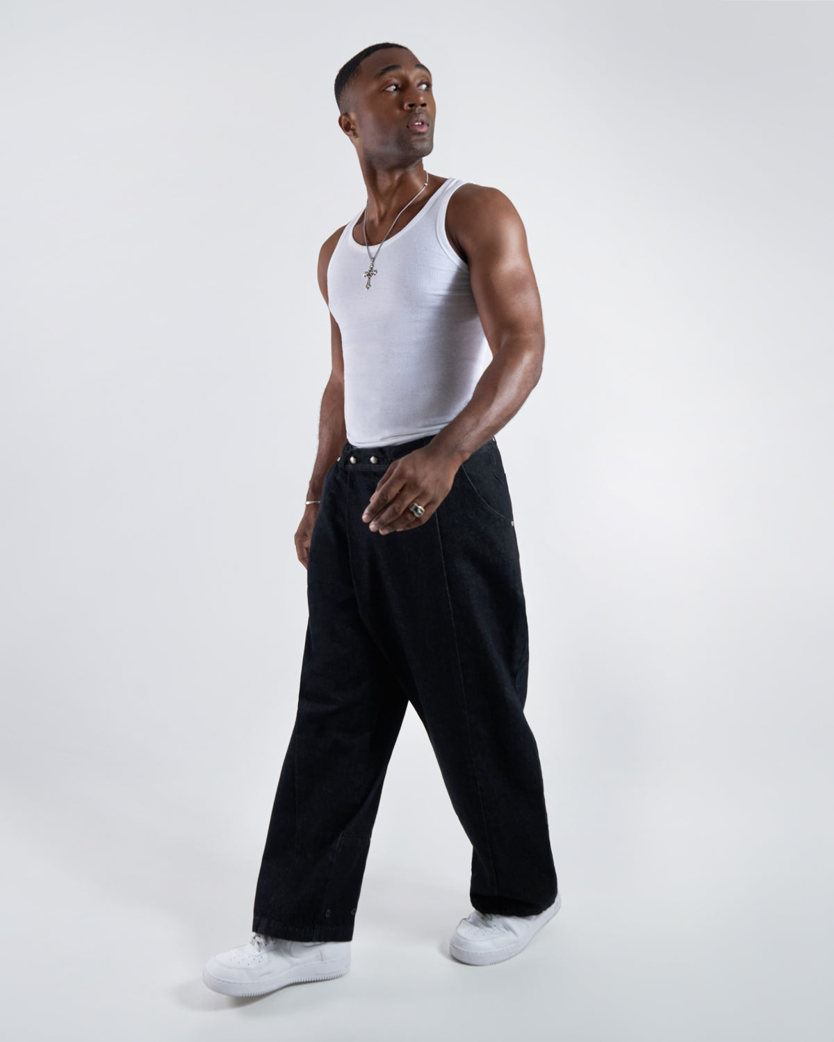 Black wide-leg hanbok denim pants modeled with white ribbed tank top and silver cross necklace, dynamic movement pose with white sneakers against white studio background