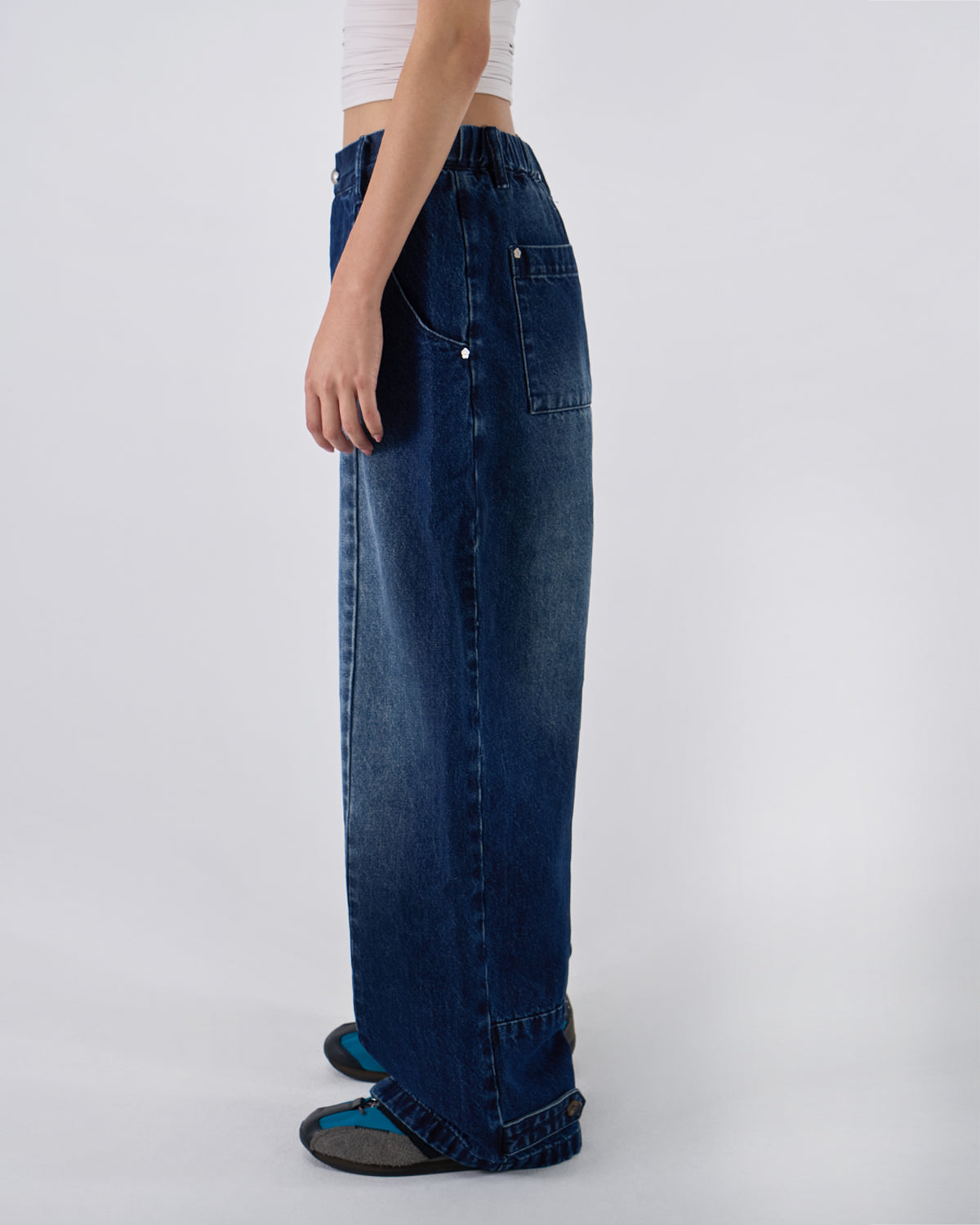 Side profile view of indigo fade wide-leg hanbok denim pants modeled with white ruched crop top, hand in front pocket, showing relaxed fit and wide-leg silhouette against white studio background