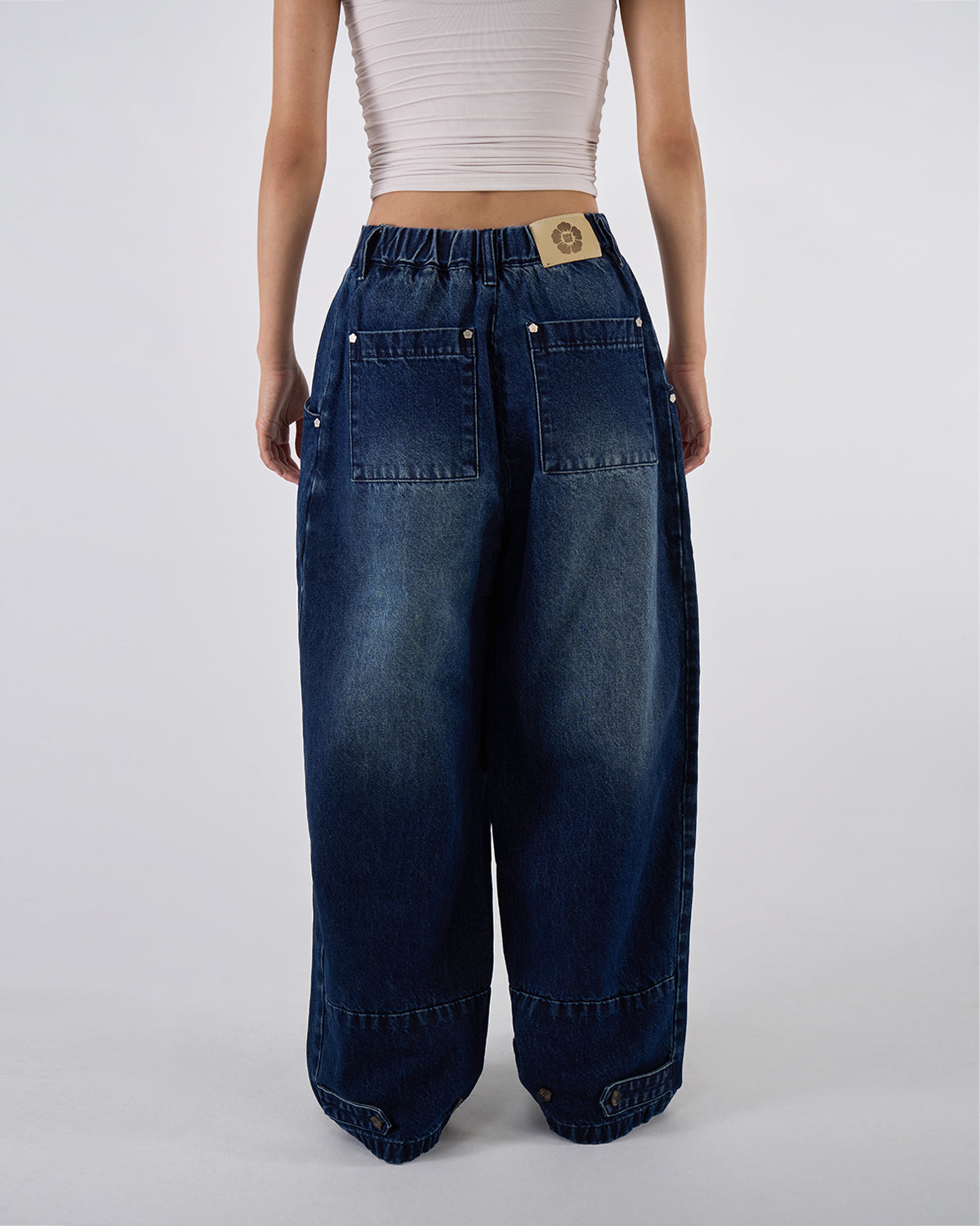 Back view of indigo fade wide-leg hanbok denim pants modeled with white ruched bandeau top, hands on waistband showing Sundae School leather patch and back pocket details against white background