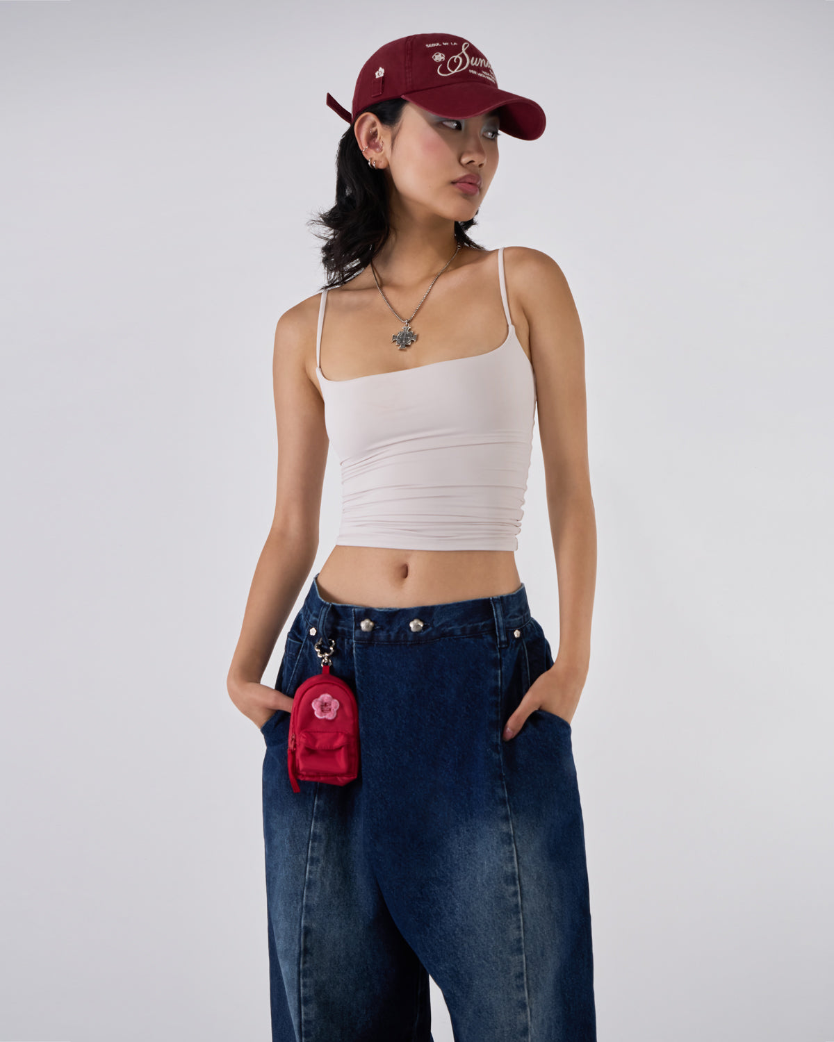 Indigo fade wide-leg hanbok denim pants modeled with white ruched bandeau top, burgundy Sundae School baseball cap, silver cross necklace, and red mini backpack charm, hands in pockets pose against white studio background