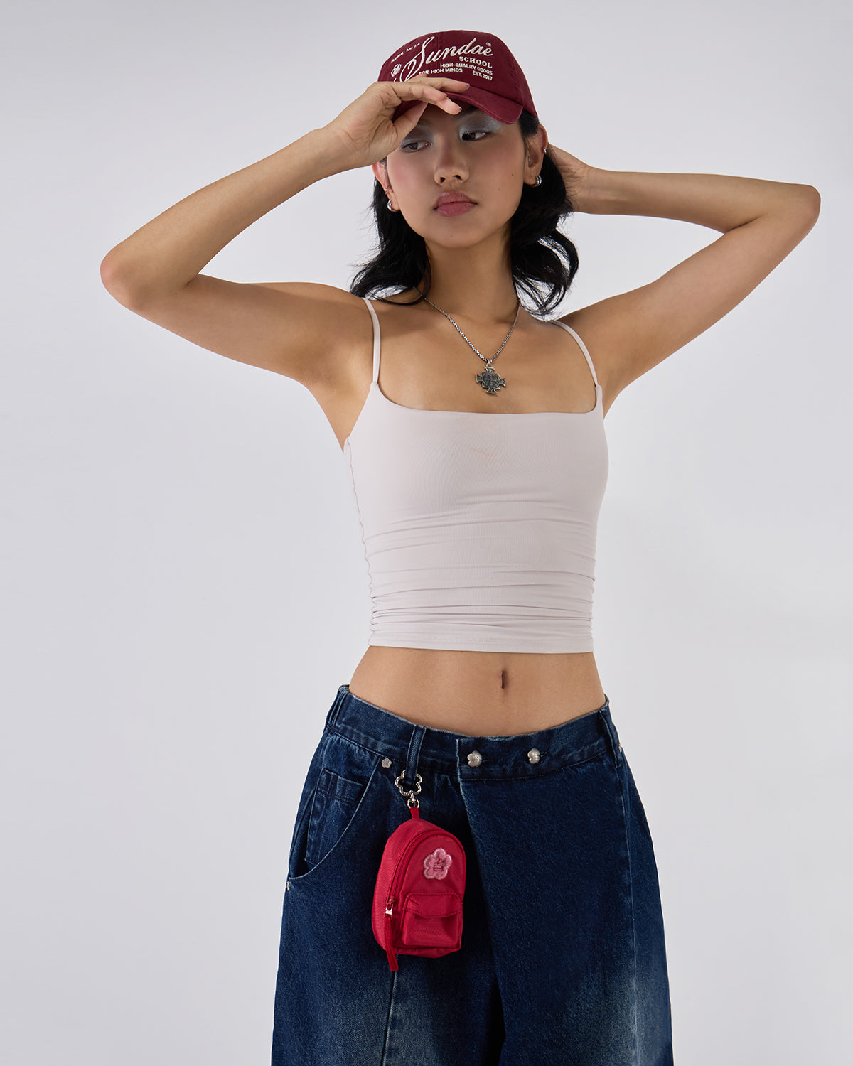 Indigo fade wide-leg hanbok denim pants modeled with white ruched bandeau top and burgundy Sundae School cap, hands raised behind head pose, silver cross necklace and red mini backpack visible against white background