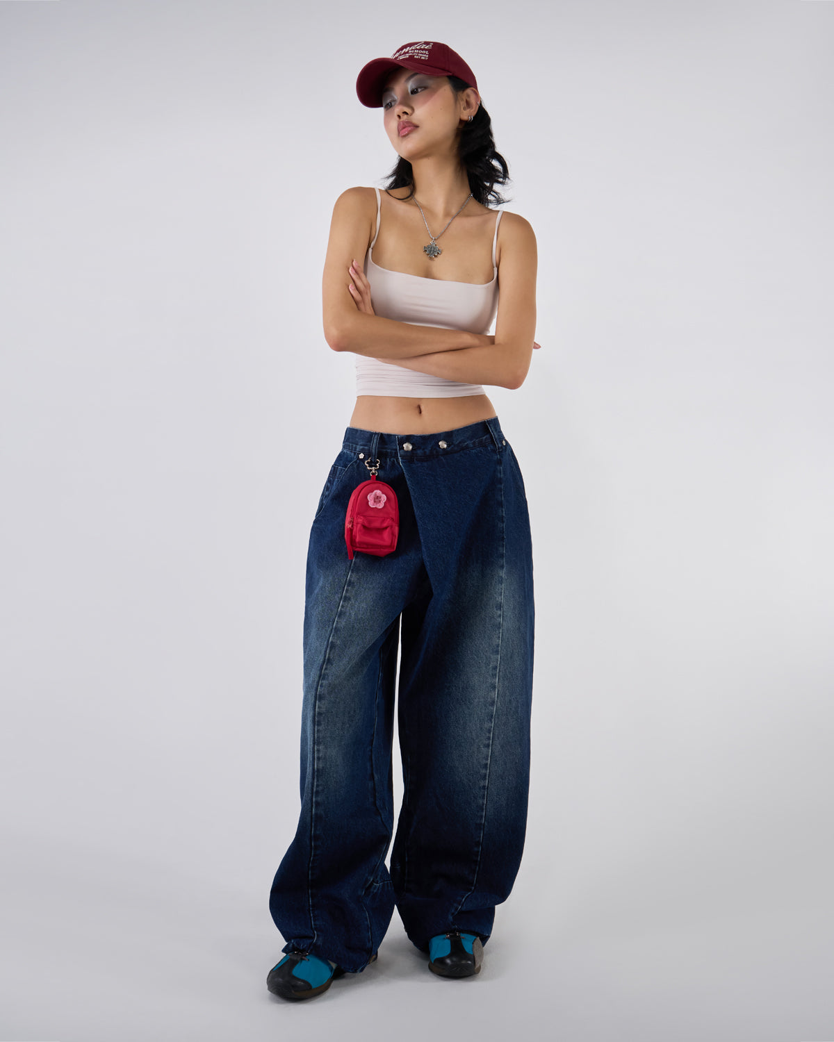 Indigo fade wide-leg hanbok denim pants modeled with white ruched bandeau top and burgundy Sundae School baseball cap, arms crossed pose, silver cross necklace and red mini backpack charm against white studio background