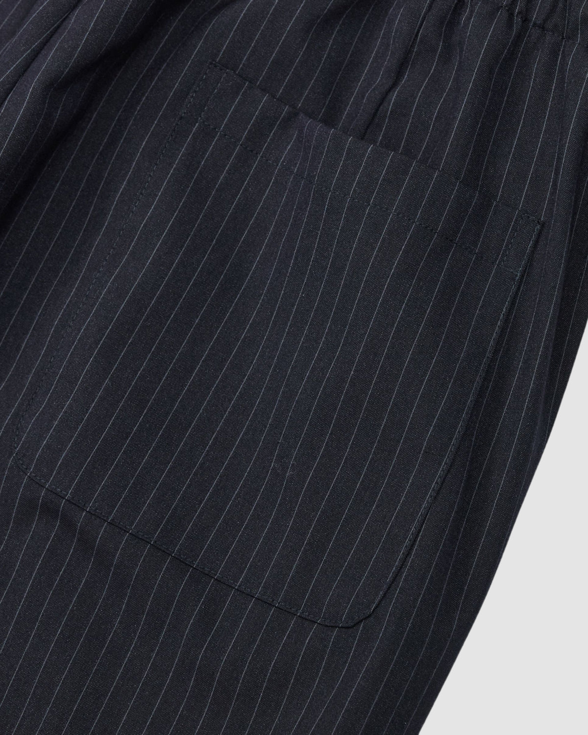Close-up of black fabric with thin white pinstripes and a sewn-on pocket from Sundae School's KIKI DRAWSTRING SLACKS. These sweatpants feature a relaxed barrel leg and an elastic drawstring waist for comfort.