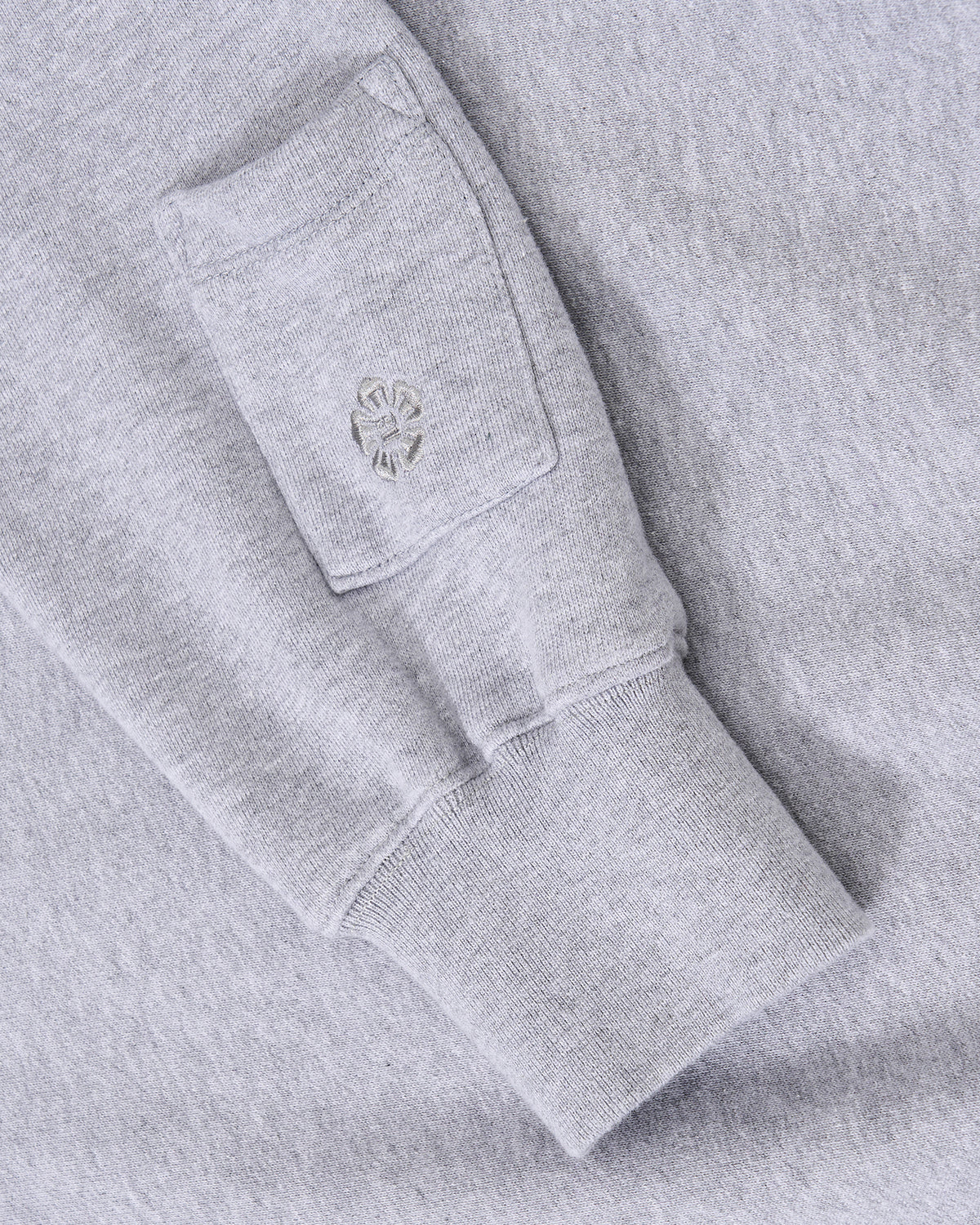 A close-up of the sleeve of the Sundae School HEATHER GREY KNIT TIE HOODIE, crafted from soft brushed cotton, featuring a small rectangular pocket with an embroidered design above the cuff.