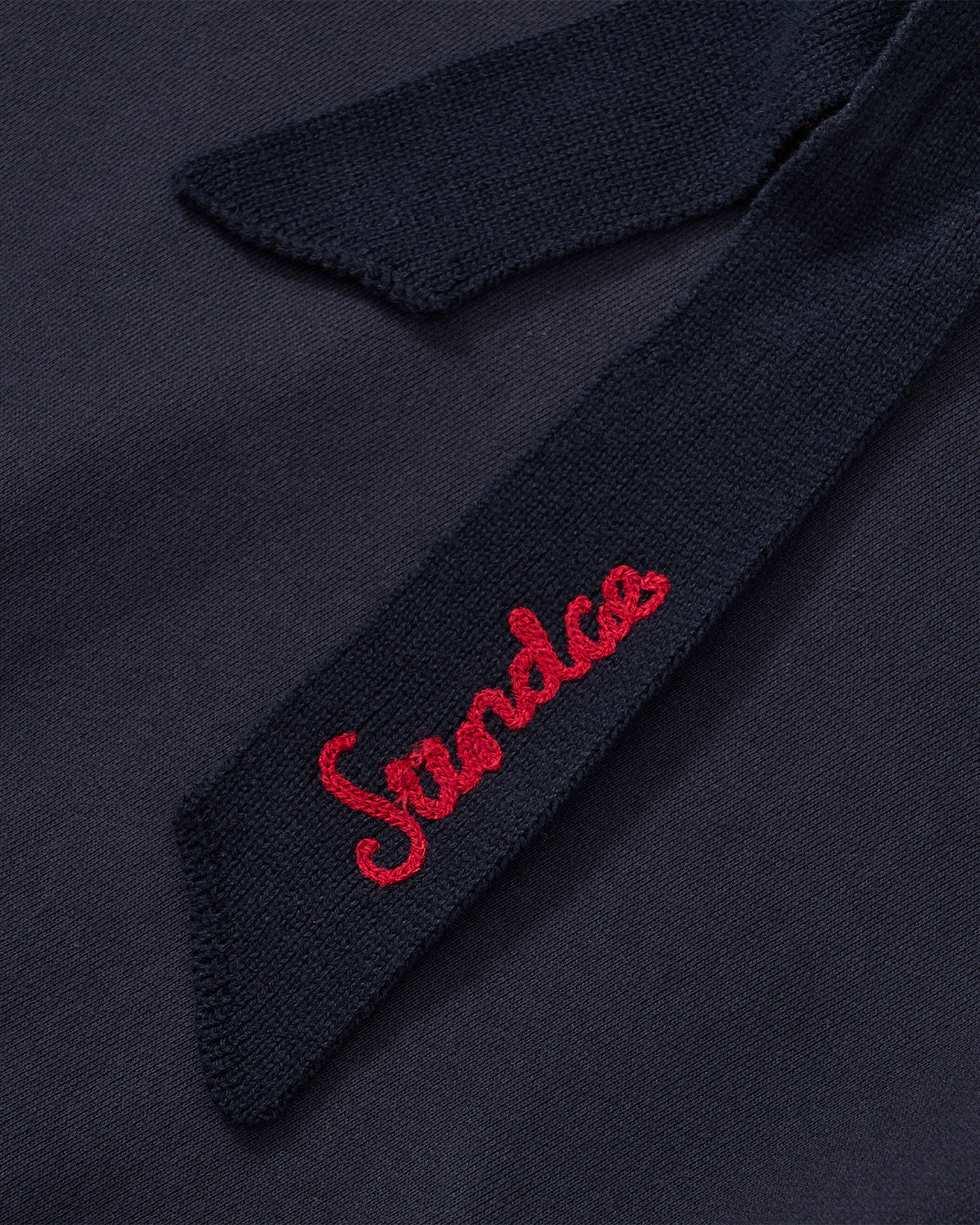 The NAVY KNIT TIE HOODIE from Sundae School features a navy blue brushed cotton fabric and a coordinating knit tie, with “Sundco” embroidered in red cursive on the tie. Designed for a unisex fit, it delivers cozy comfort with its heavyweight construction.