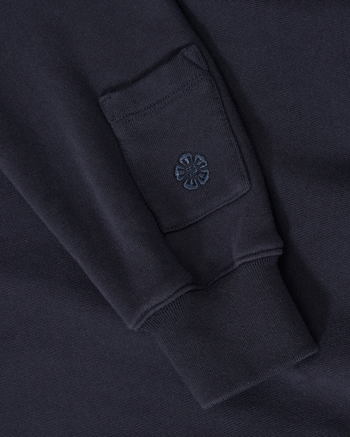 Close-up of the Sundae School NAVY KNIT TIE HOODIE sleeve, featuring a small square patch pocket and an embroidered flower design near the cuff. The unisex fit and brushed cotton provide comfort and style to this standout piece.