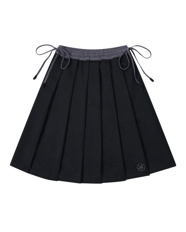 Introducing the BLACK KONA MIDI SKIRT by Sundae School—a black pleated skirt featuring a gray waistband and adjustable side ties. A small embroidered flower accent decorates one corner. The skirt is displayed laid flat against a white background.