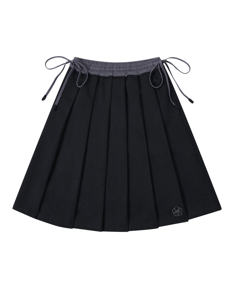 Introducing the BLACK KONA MIDI SKIRT by Sundae School—a black pleated skirt featuring a gray waistband and adjustable side ties. A small embroidered flower accent decorates one corner. The skirt is displayed laid flat against a white background.