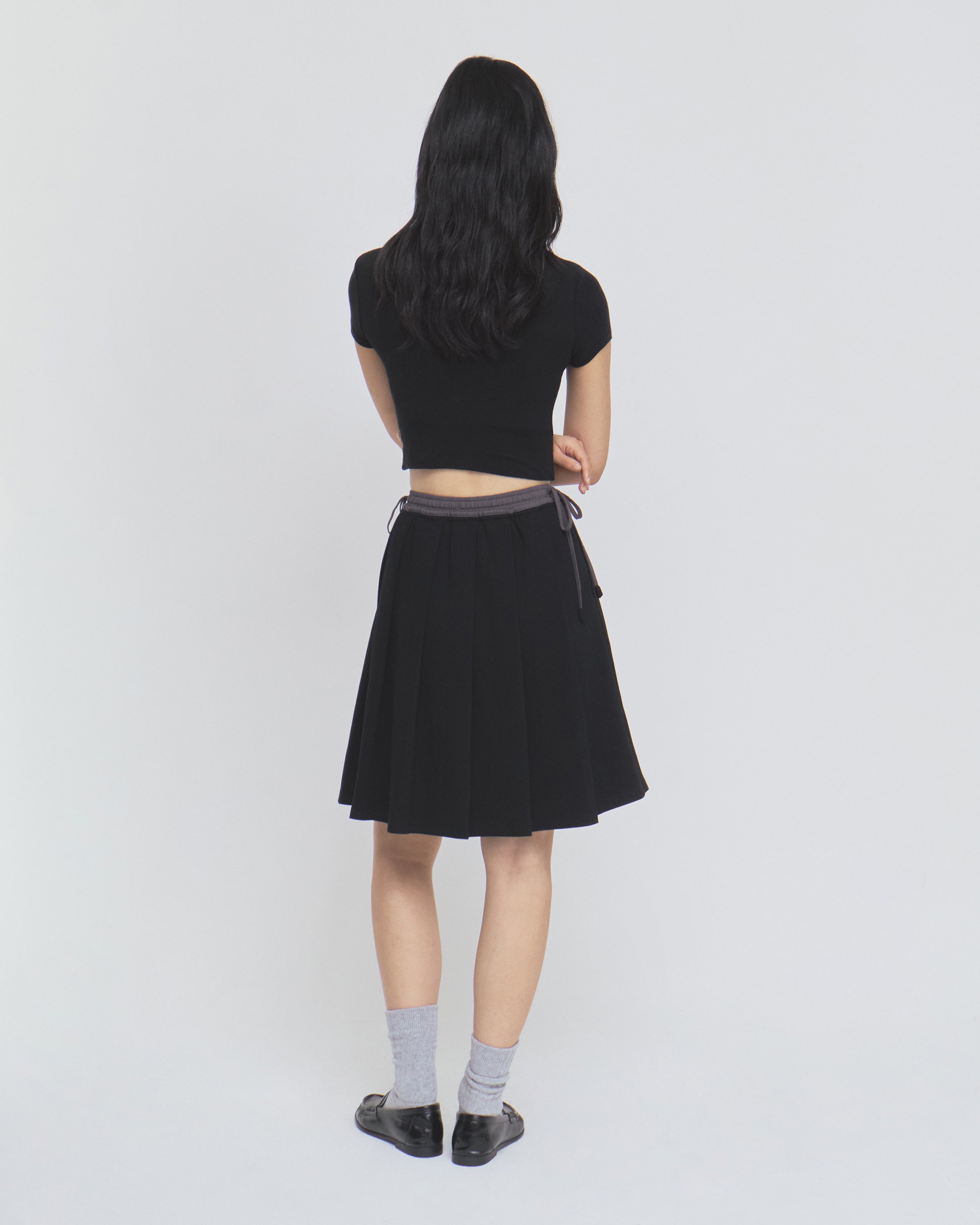 A person with long dark hair, dressed in a black shirt and the BLACK KONA MIDI SKIRT by Sundae School, along with gray socks and black shoes, stands facing away from the camera against a plain white background.