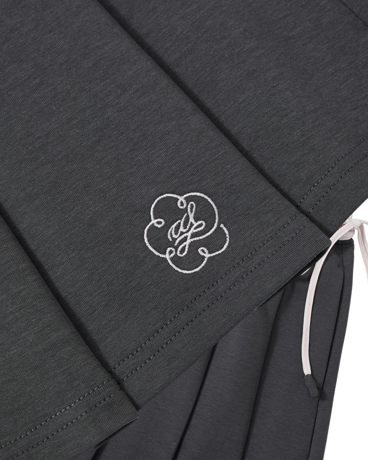 Close-up of the CHARCOAL KONA MIDI SKIRT by Sundae School, featuring dark gray pleats, an embroidered white decorative monogram, and a white ribbon detail on the right side.