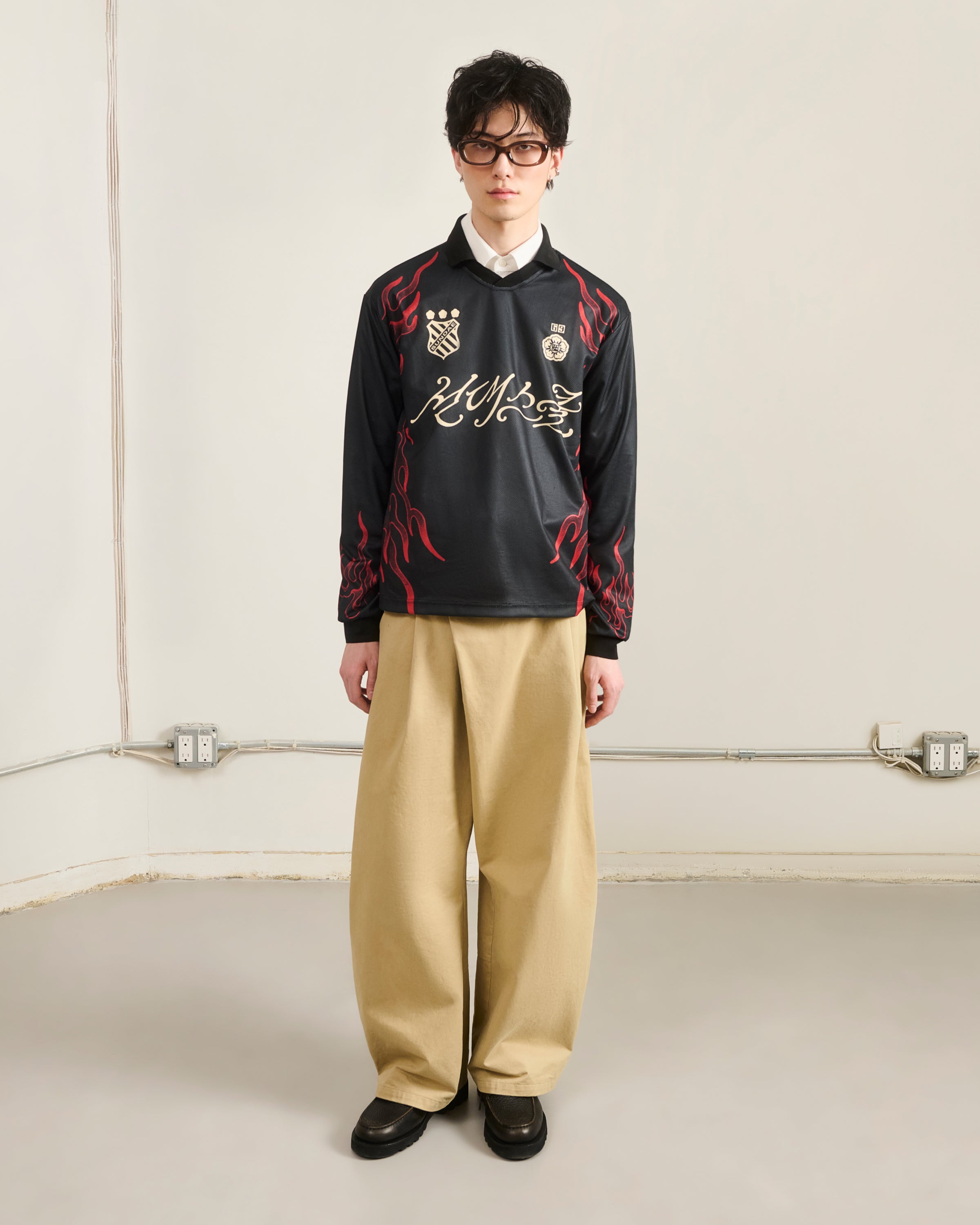 A person with short dark hair and glasses stands against a plain wall, wearing Sundae School's KOREATOWN FOOTBALL JERSEY—featuring custom Korean graphics and red flames—paired with wide beige pants and black shoes, channeling K-Town fashion.