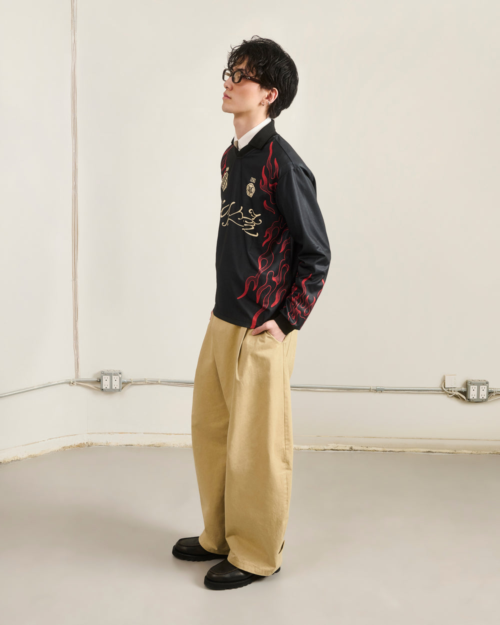 A person stands sideways in a plain room, hands in pockets, wearing Sundae School’s KOREATOWN FOOTBALL JERSEY with red Korean-inspired flame designs, beige wide-leg pants, black shoes, and dark glasses.