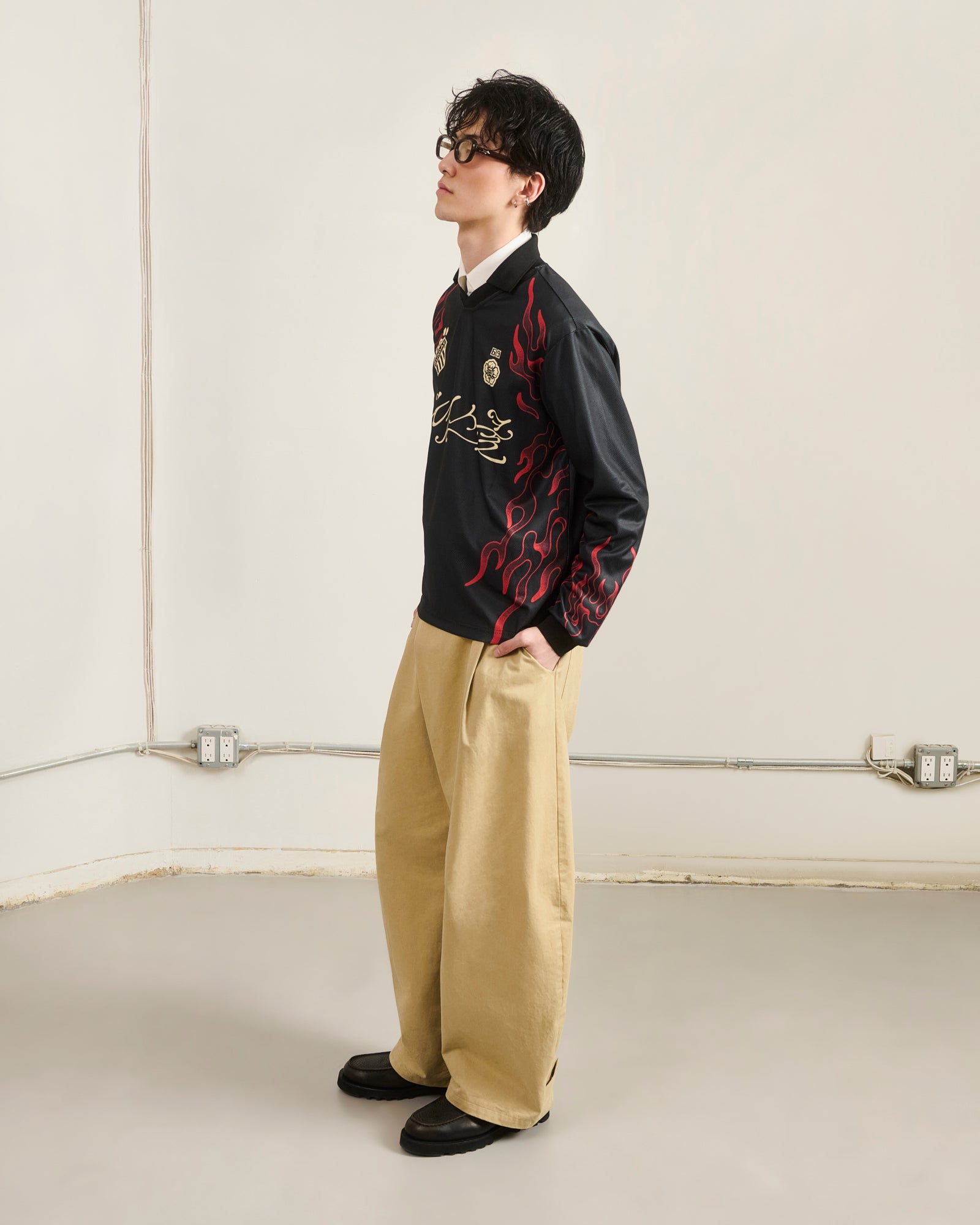 A person stands sideways in a plain room, hands in pockets, wearing Sundae School’s KOREATOWN FOOTBALL JERSEY with red Korean-inspired flame designs, beige wide-leg pants, black shoes, and dark glasses.