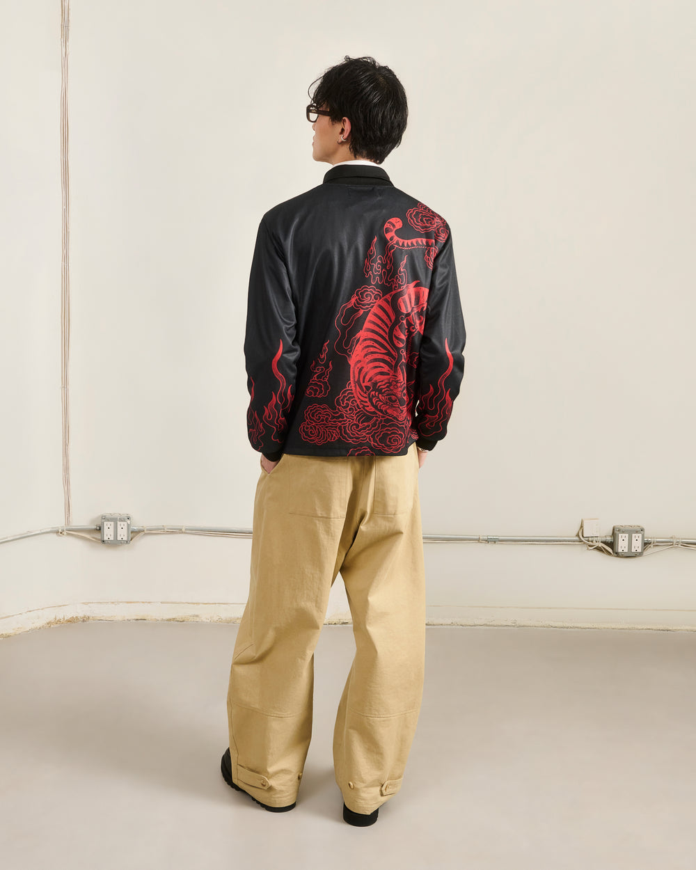 A person stands facing away, wearing a black Sundae School KOREATOWN FOOTBALL JERSEY with a red dragon design on the back and loose beige pants, in a minimal room with white walls and visible electrical outlets.
