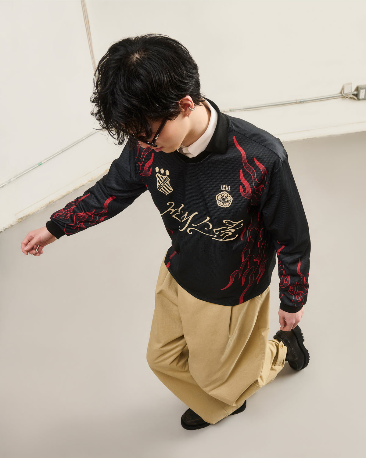 A person with short dark hair, glasses, and loose beige pants kneels indoors on a light floor wearing Sundae School’s KOREATOWN FOOTBALL JERSEY—a black top with red flame and custom Korean graphics.