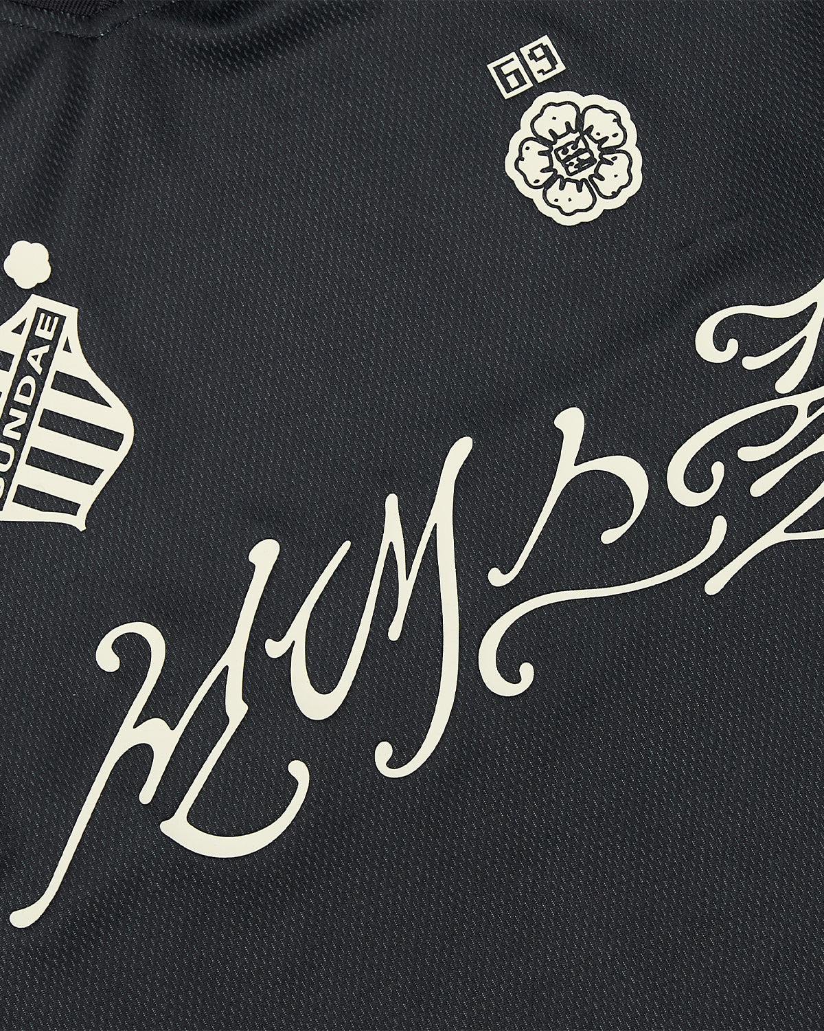 Close-up of a black KOREATOWN FOOTBALL JERSEY by Sundae School, featuring white symbols: a crest with “SUNDAE,” a flower marked with 69, and decorative calligraphy across the center—perfect for fans of custom script apparel and distinctive K-Town fashion.