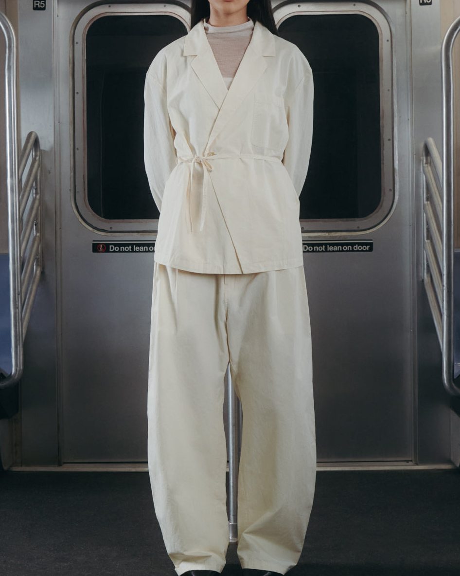 A person wearing the IVORY LINEN GORUM BLAZER and IVORY FLOWER NORIGAE LINEN TROUSERS by Sundae School stands in the center of an empty subway car, hands behind their back and facing forward. The subway features silver walls and blue seats.