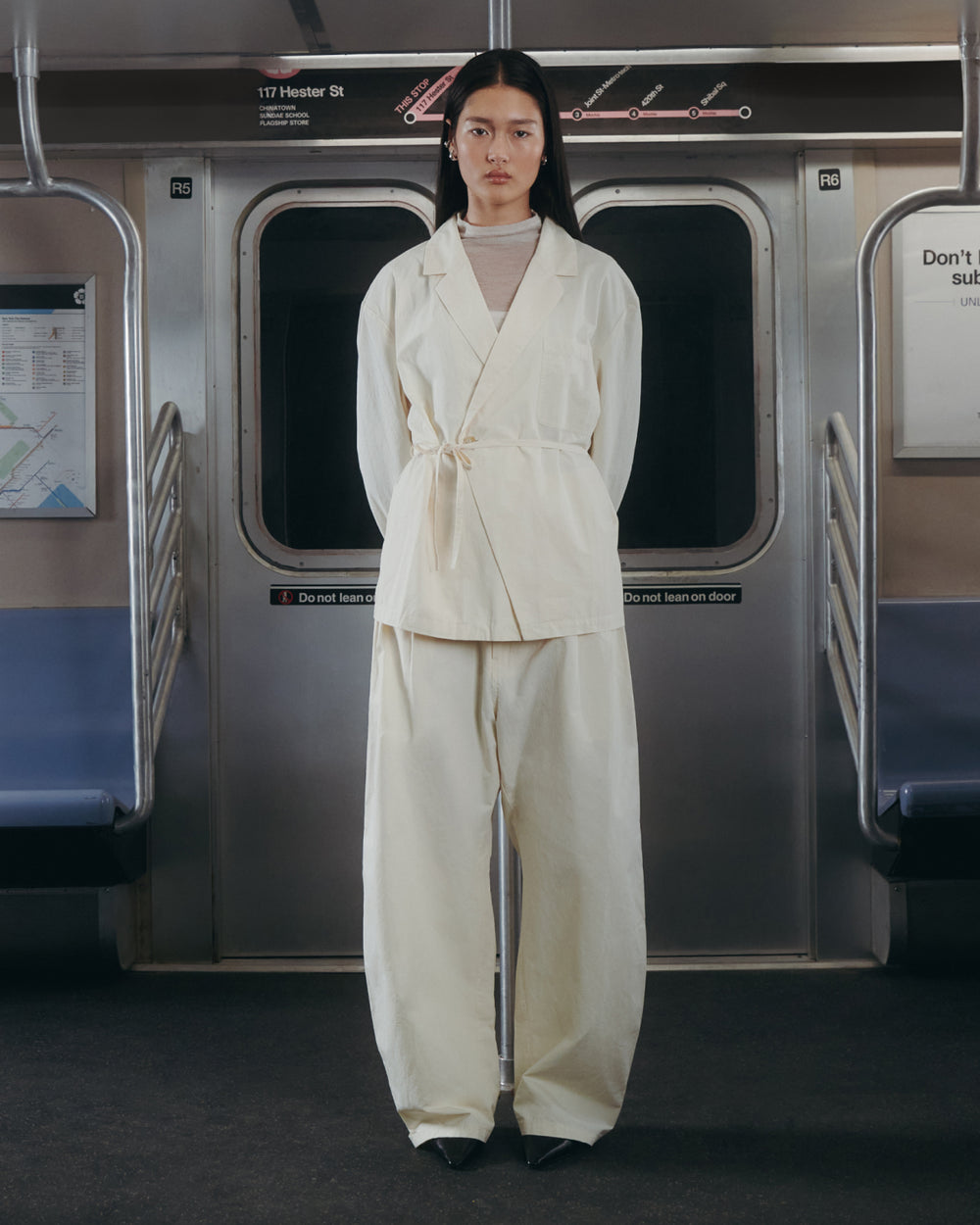 A person wearing the IVORY LINEN GORUM BLAZER by Sundae School stands in the center of an empty subway car, hands behind their back and facing forward. The subway features silver walls and blue seats.