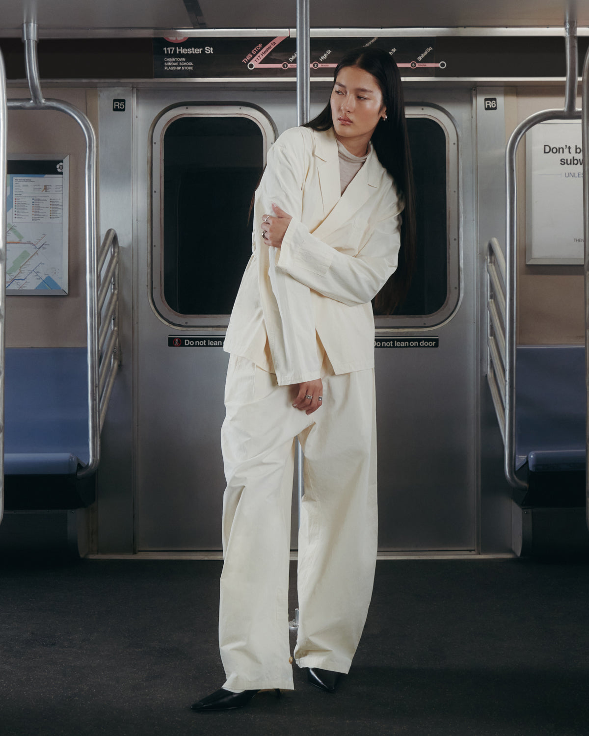 A person wearing Sundae School's IVORY FLOWER NORIGAE LINEN TROUSERS and black shoes stands in a subway car, posing with arms crossed. Subway maps and warning signs are visible on the walls behind them.