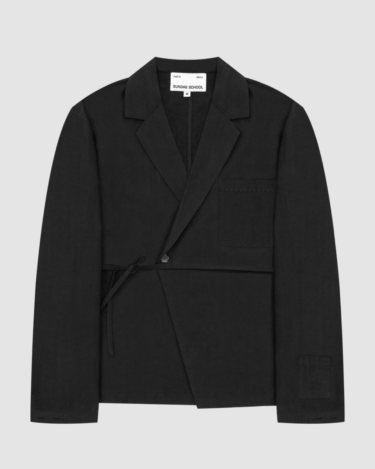 The BLACK LINEN GORUM BLAZER by Sundae School features a wrap-front design, single button closure, tie detail on the left side, three pockets, and a white label on the inside collar reading SUNDAE SCHOOL. The blazer is displayed against a plain background.