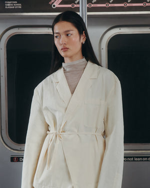 A woman stands in a subway car, wearing the IVORY LINEN GORUM BLAZER by Sundae School over a beige turtleneck. She has long, straight dark hair and a neutral expression.