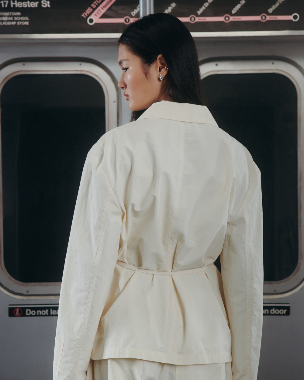 A woman with long dark hair, dressed in the Sundae School IVORY LINEN GORUM BLAZER, stands in front of subway doors, facing away and glancing to the side, with subway maps and signs displayed above.