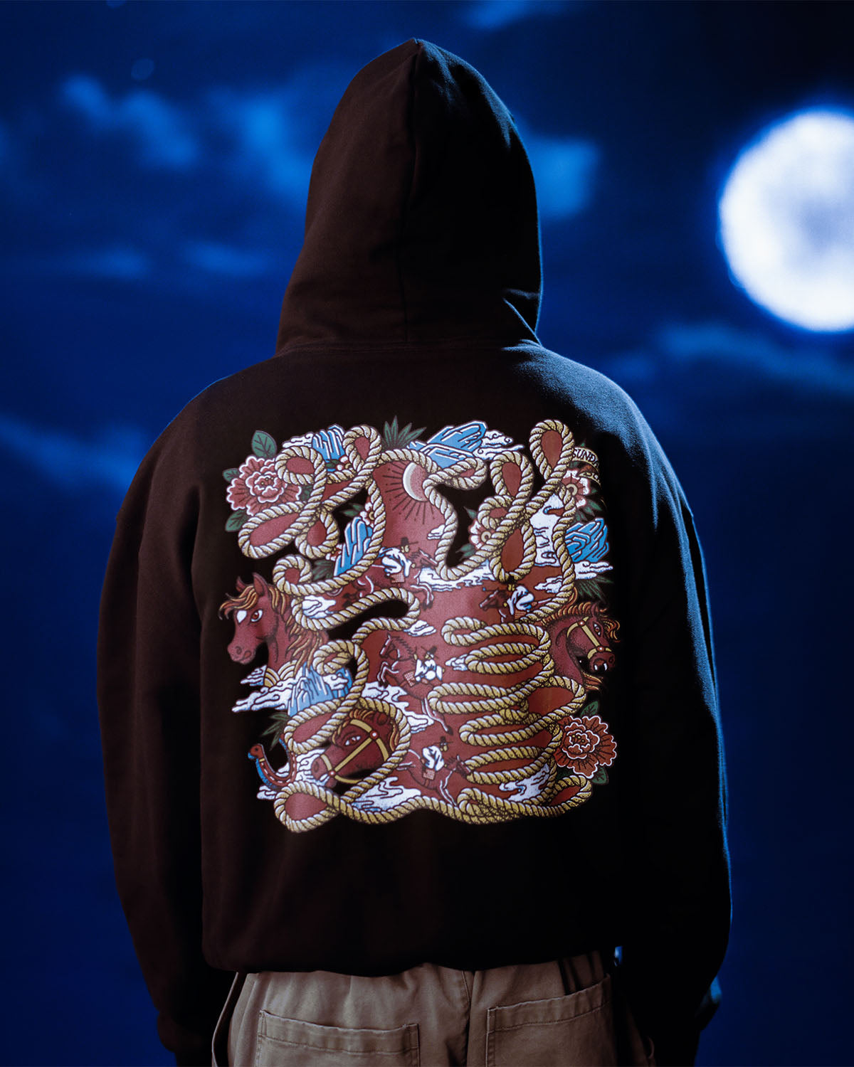 A person in the Sundae School HIGH HORSE LNY HOODIE, featuring a vibrant back graphic of snakes, a bull, flowers, and ropes, stands under a cloudy night sky illuminated by a bright full moon.