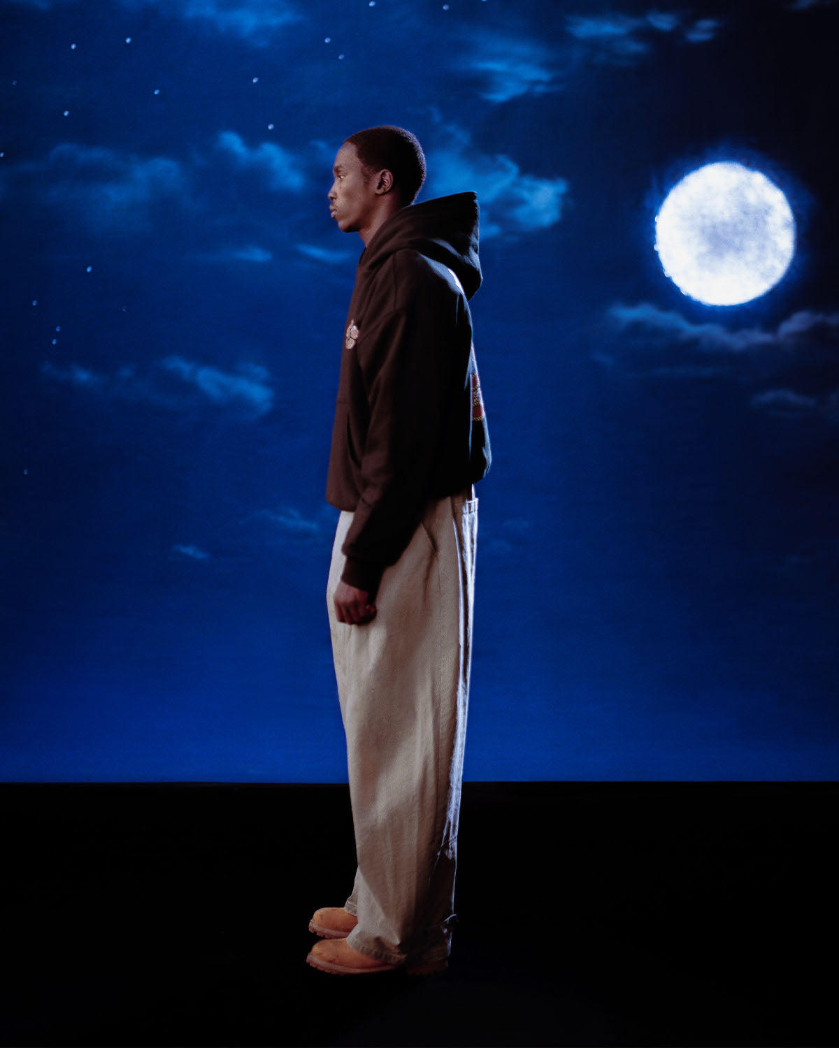 Wearing the Sundae School HIGH HORSE LNY HOODIE and light pants, a person stands in profile before a night sky with a glowing full moon and scattered stars.