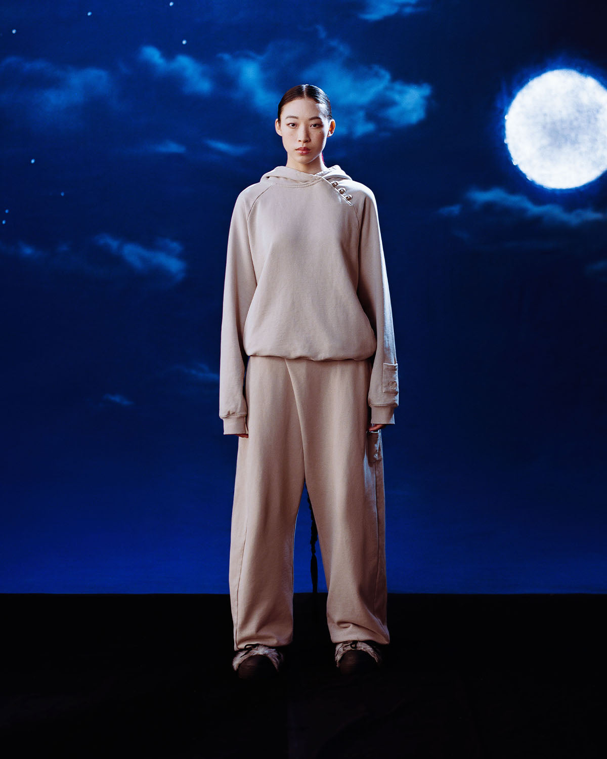 A person wearing the TAUPE WRAP SWEATBAJI by Sundae School stands against a dark blue night sky filled with clouds and a bright full moon.