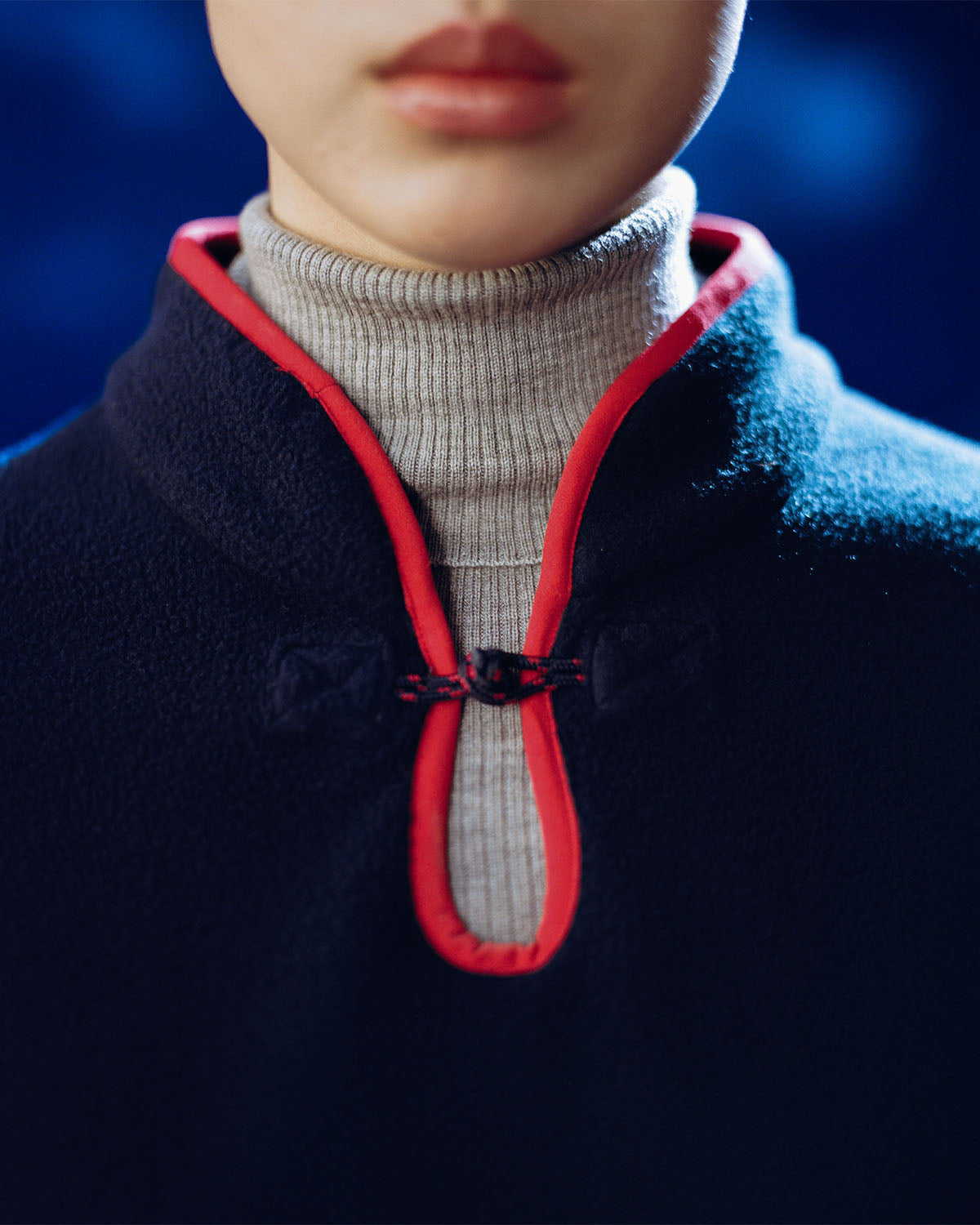A close-up of a person wearing the Sundae School NAVY TEARDROP KNOT FLEECE, featuring red trim and a frog closure over a light gray ribbed turtleneck. Only the lower face and neck show against a blurred blue background.