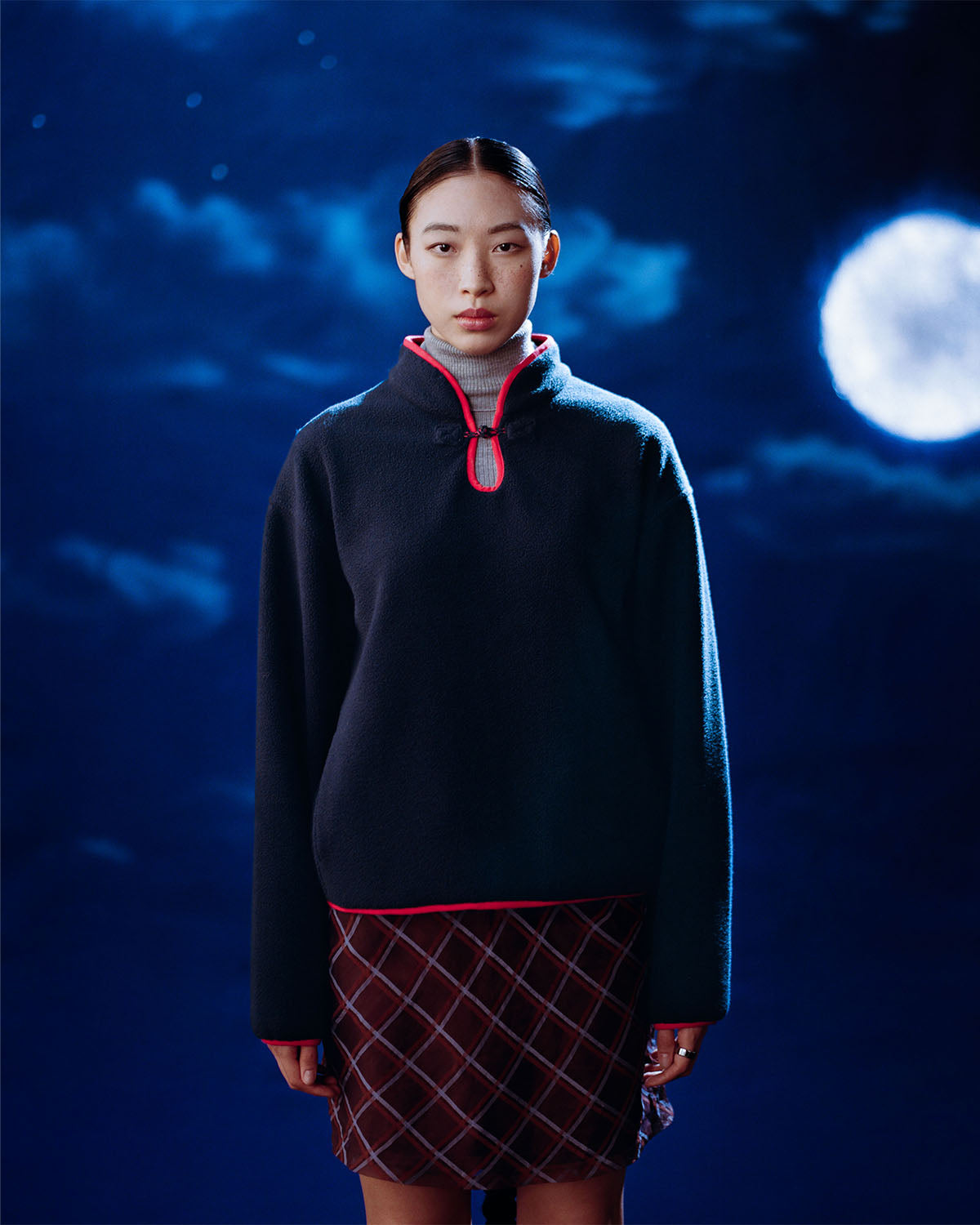 A young woman stands against a dark blue night sky with clouds and a full moon, calmly facing the camera in the Sundae School NAVY TEARDROP KNOT FLEECE.