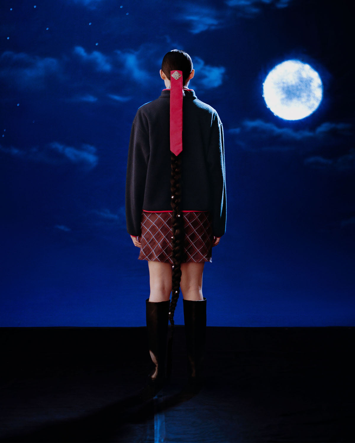 A person with long braided hair tied with a red ribbon stands before a full moon, wearing the Sundae School NAVY TEARDROP KNOT FLEECE, patterned skirt, and knee-high boots amid a dreamy, surreal night sky.