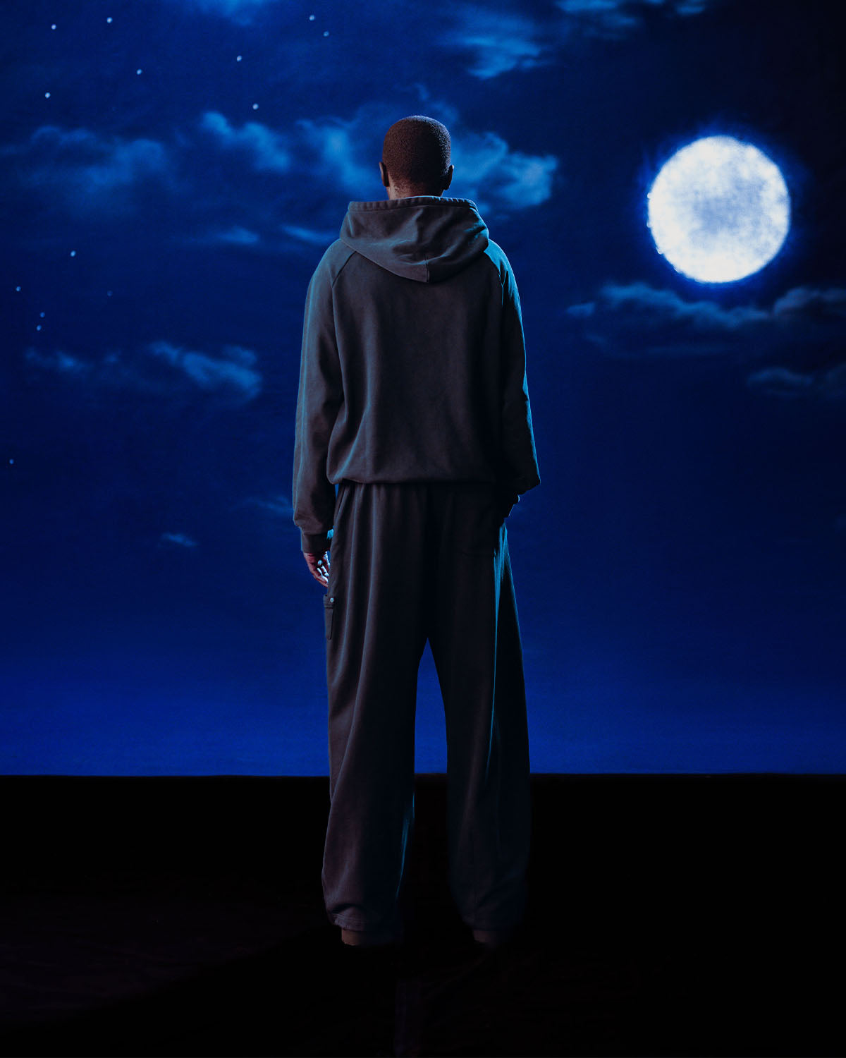 A man stands before a moon, wearing Sundae School’s INDIGO WASH HANBOK HOODIE featuring a custom 