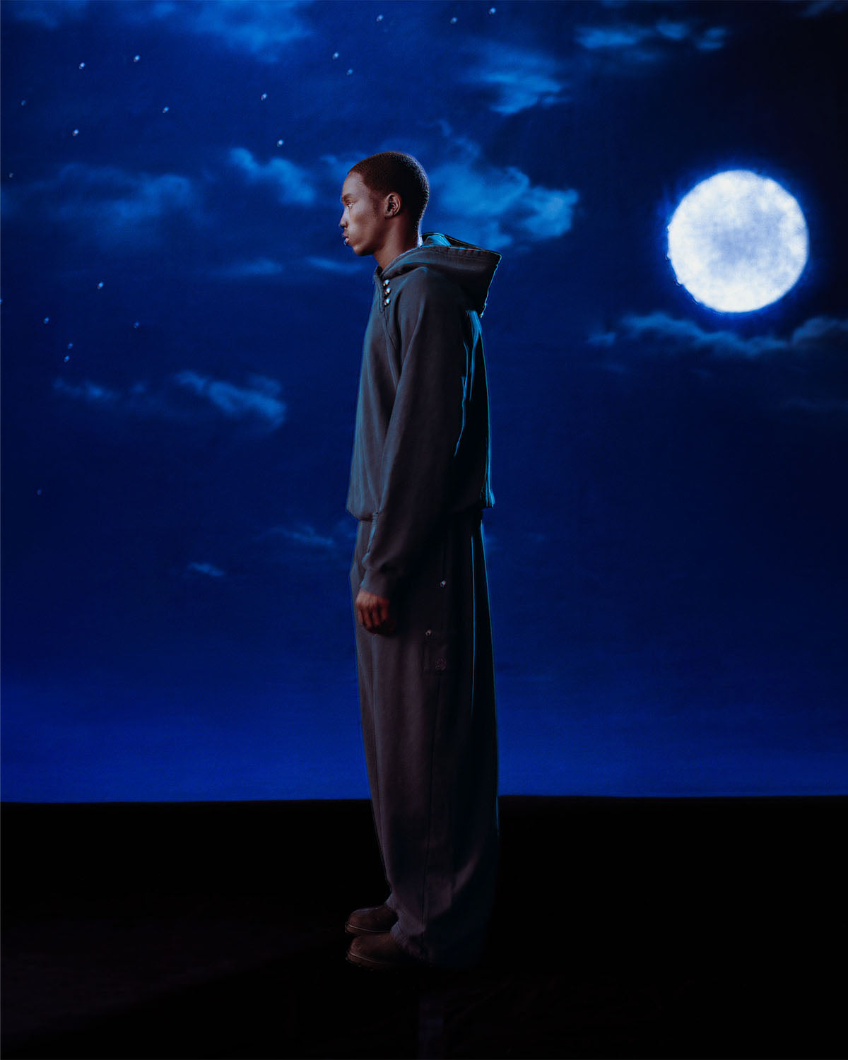 A person in loose, dark clothing stands in profile against a night sky, lit by a full moon, wearing Sundae School's INDIGO WASH HANBOK HOODIE featuring a custom 