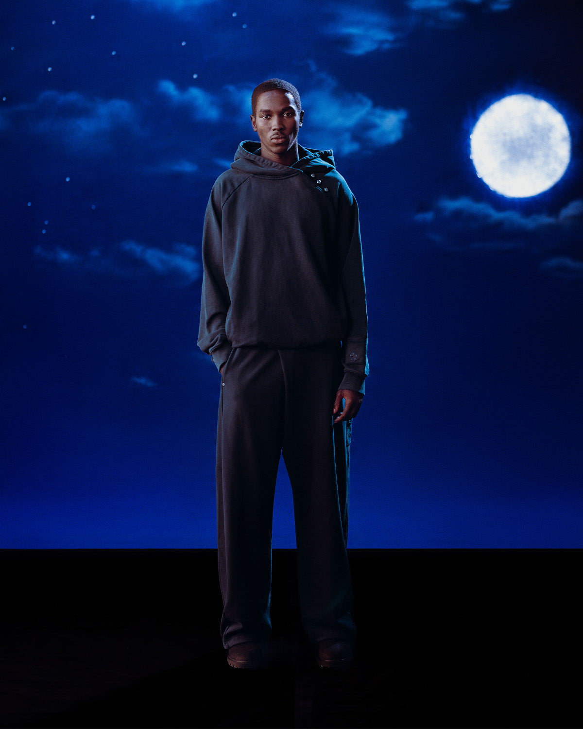 A man stands calmly against a deep blue night sky, wearing Sundae School’s INDIGO WASH HANBOK HOODIE with its signature sleeve stash pocket. He faces forward, hands at his sides, dressed in loose, dark clothing beneath the bright full moon and clouds.
