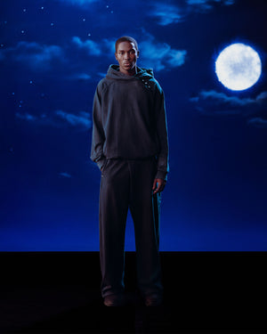 A man stands calmly against a deep blue night sky, wearing Sundae School’s INDIGO WASH HANBOK HOODIE with its signature sleeve stash pocket. He faces forward, hands at his sides, dressed in loose, dark clothing beneath the bright full moon and clouds.
