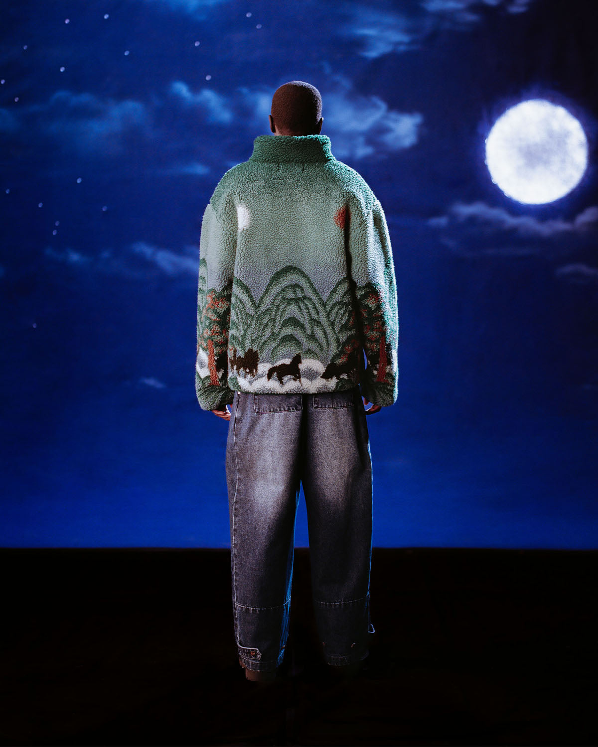 A person stands with their back to the camera, wearing the Sundae School TEAL FIVE PEAKS FLEECE—featuring a mountain and animal design—and wide-leg jeans, set against a dark backdrop with a full moon and clouds.