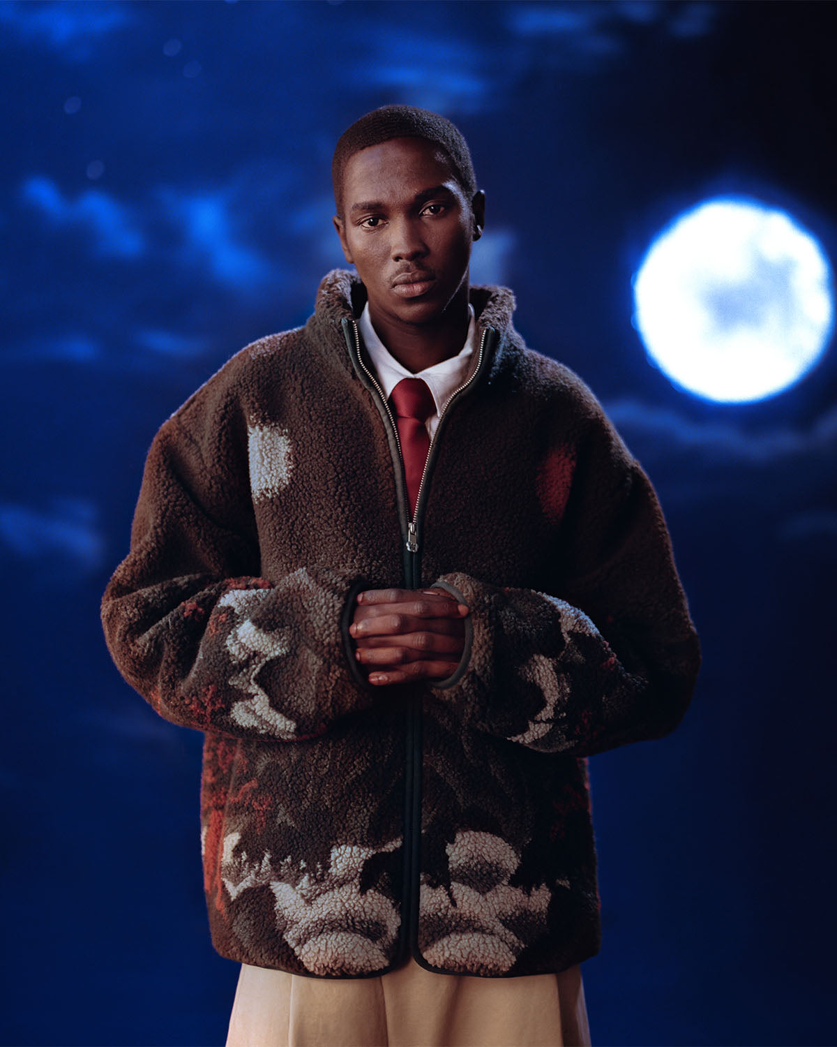 A man in a Sundae School CHARCOAL FIVE PEAKS FLEECE, white shirt, and red tie stands with hands clasped against a night sky backdrop featuring a full moon and deep blue hues.