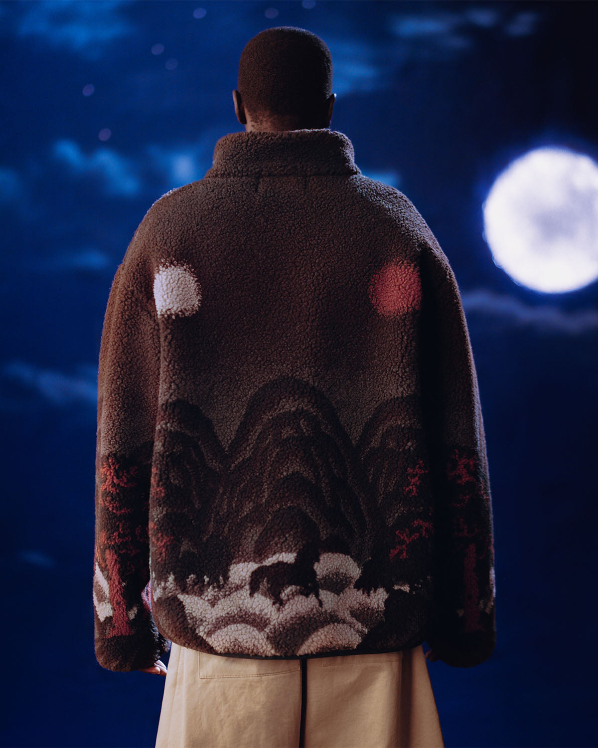 A person faces away from the camera, wearing Sundae School's CHARCOAL FIVE PEAKS FLEECE—a charcoal embossed pile fleece jacket with mountains and trees beneath a full moon in a dark blue night sky.