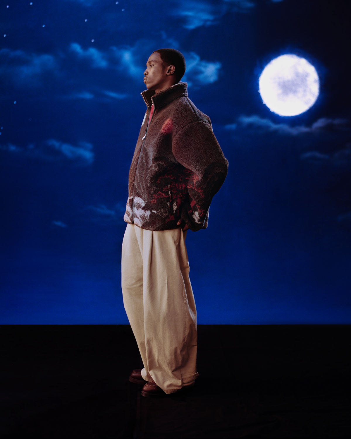 A person stands in profile against a dark, starry night sky with a bright full moon, hands behind their back, gazing thoughtfully into the distance while wearing Sundae School’s CHARCOAL FIVE PEAKS FLEECE and loose cream pants.